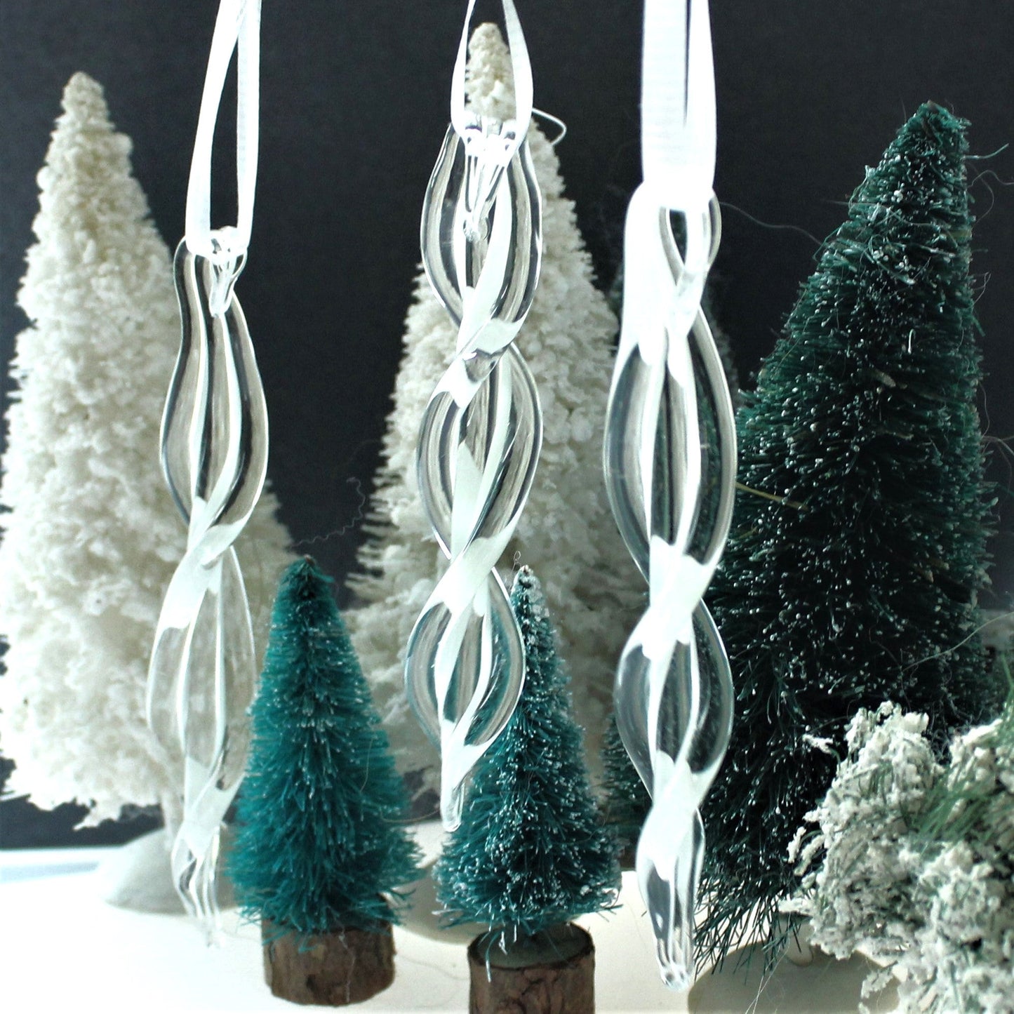 Three handmade glass ornaments with twisted spiral design displayed with miniature Christmas trees. Ideal for seasonal and holiday decorations.