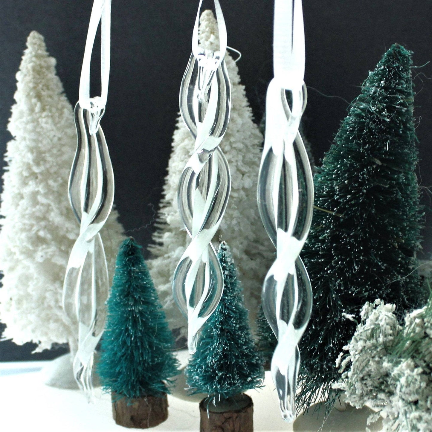 Three handmade glass ornaments with twisted spiral design displayed with miniature Christmas trees. Ideal for seasonal and holiday decorations.