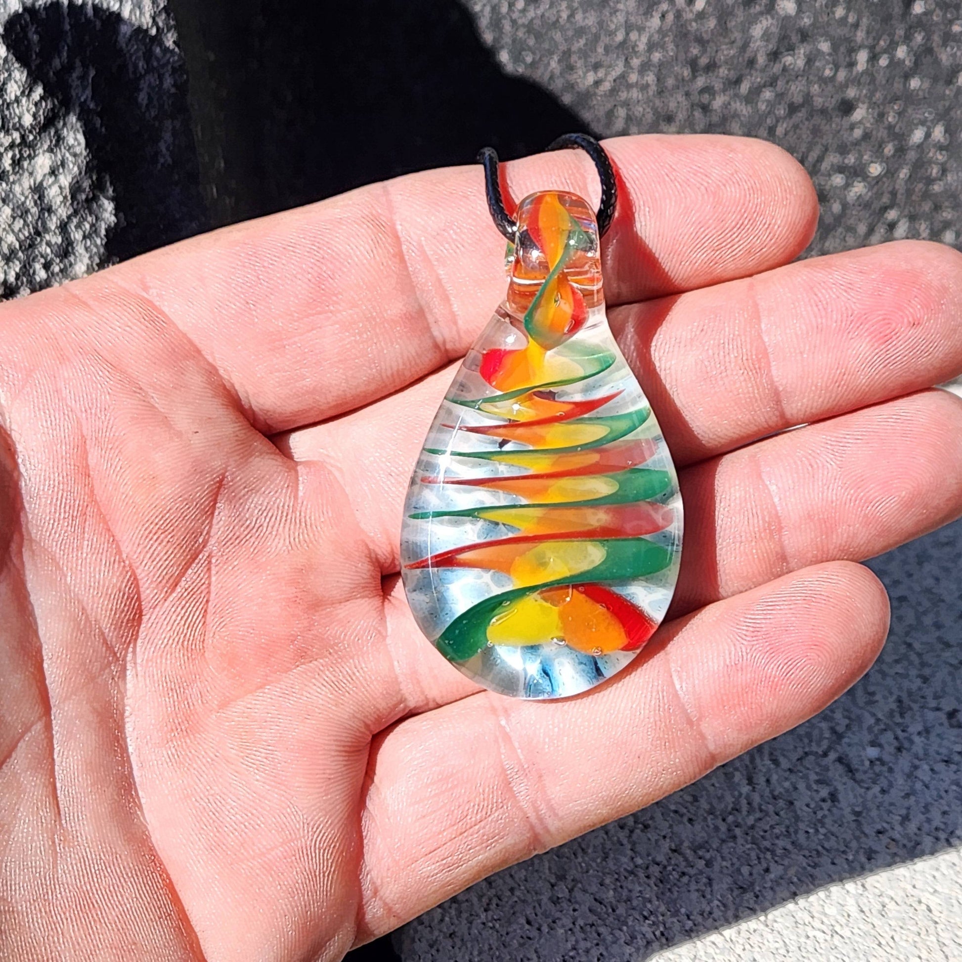 Hand holding handmade glass pendant necklace with colorful spiral rainbow design, showing the pendant's size and clarity in natural light.