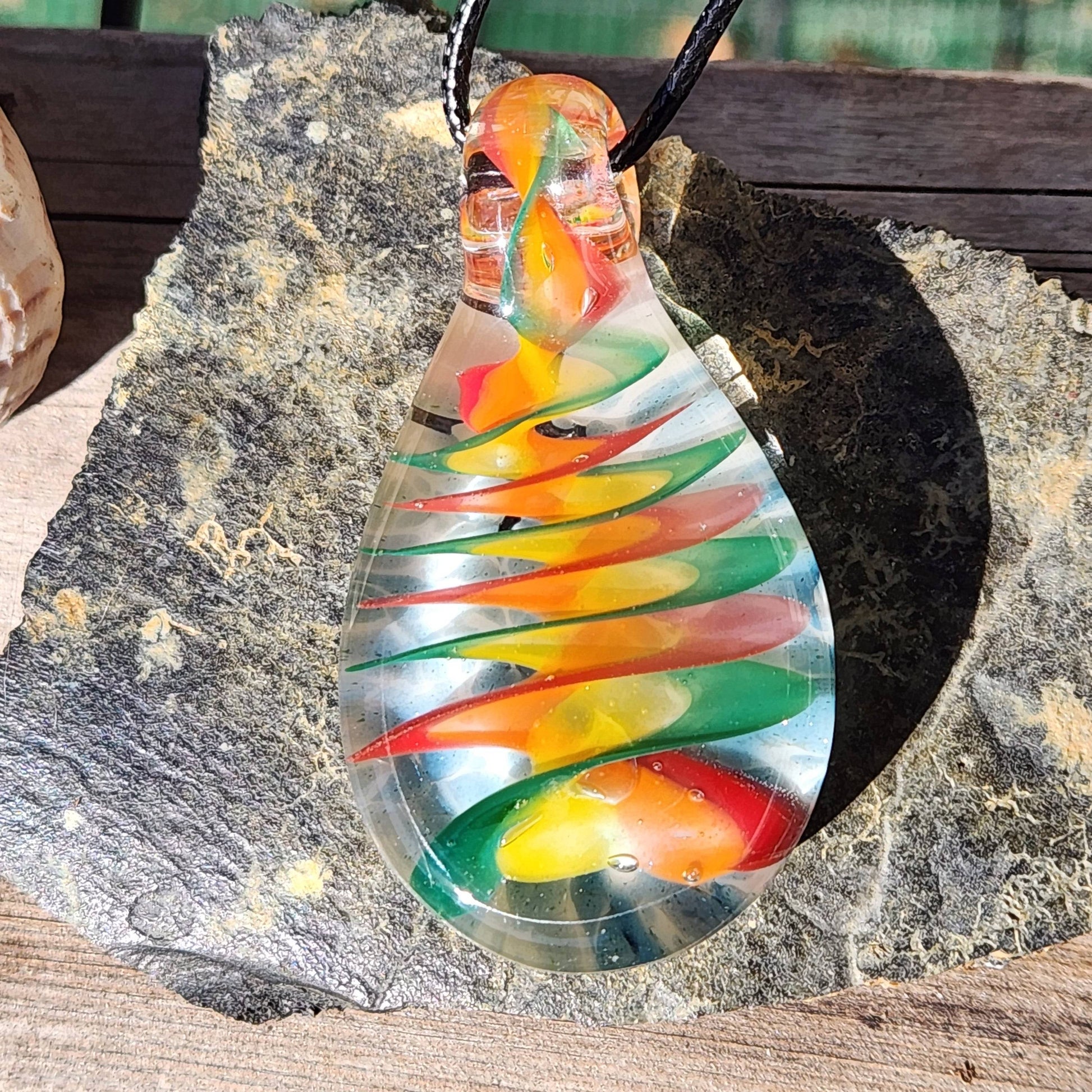 Handmade glass pendant necklace with spiral rainbow pattern displayed on a textured rock, highlighting its vibrant colors and craftsmanship.