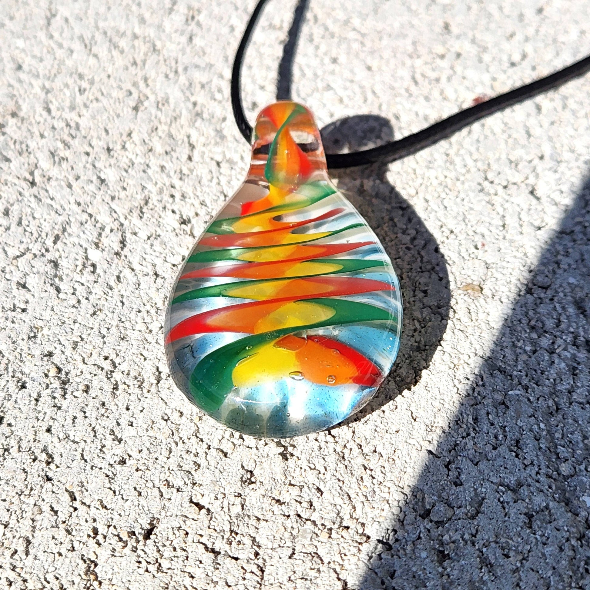 Colorful handmade glass pendant necklace with rainbow swirl pattern lying flat on a textured stone surface in bright sunlight.
