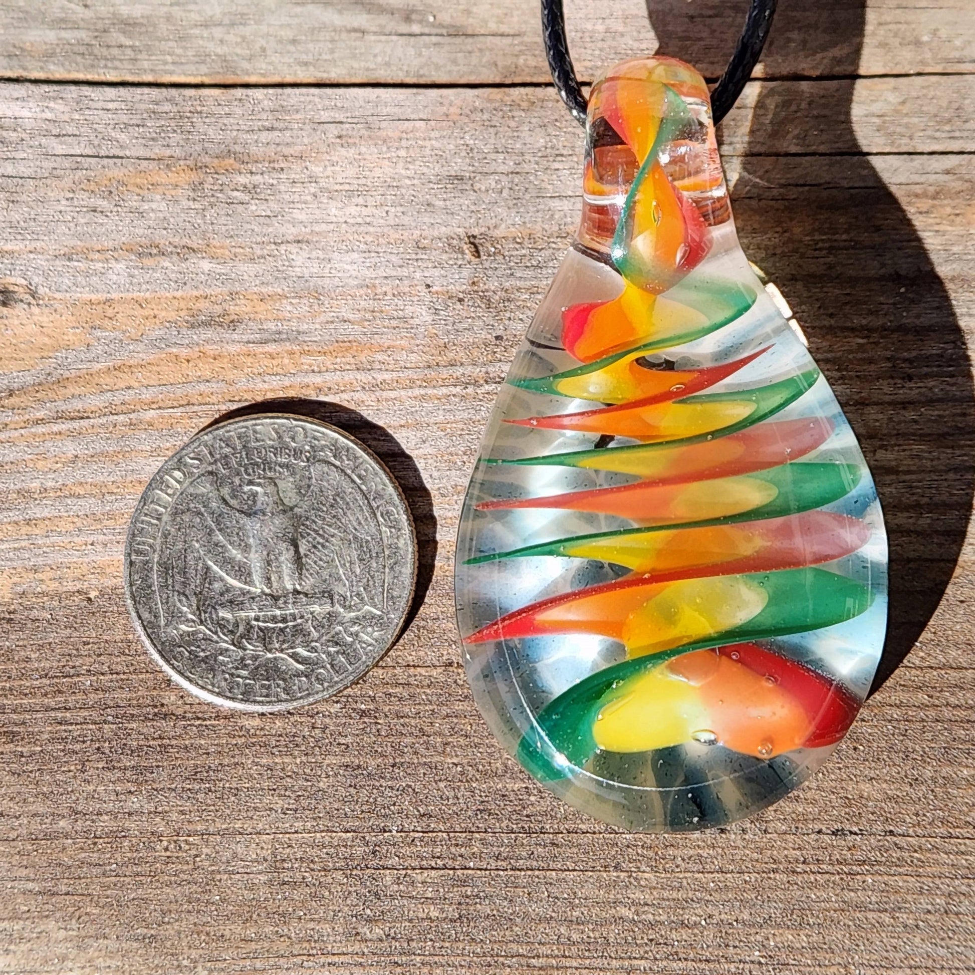 Handmade glass pendant necklace with rainbow swirl design shown next to a U.S. quarter for size comparison on a wooden surface.