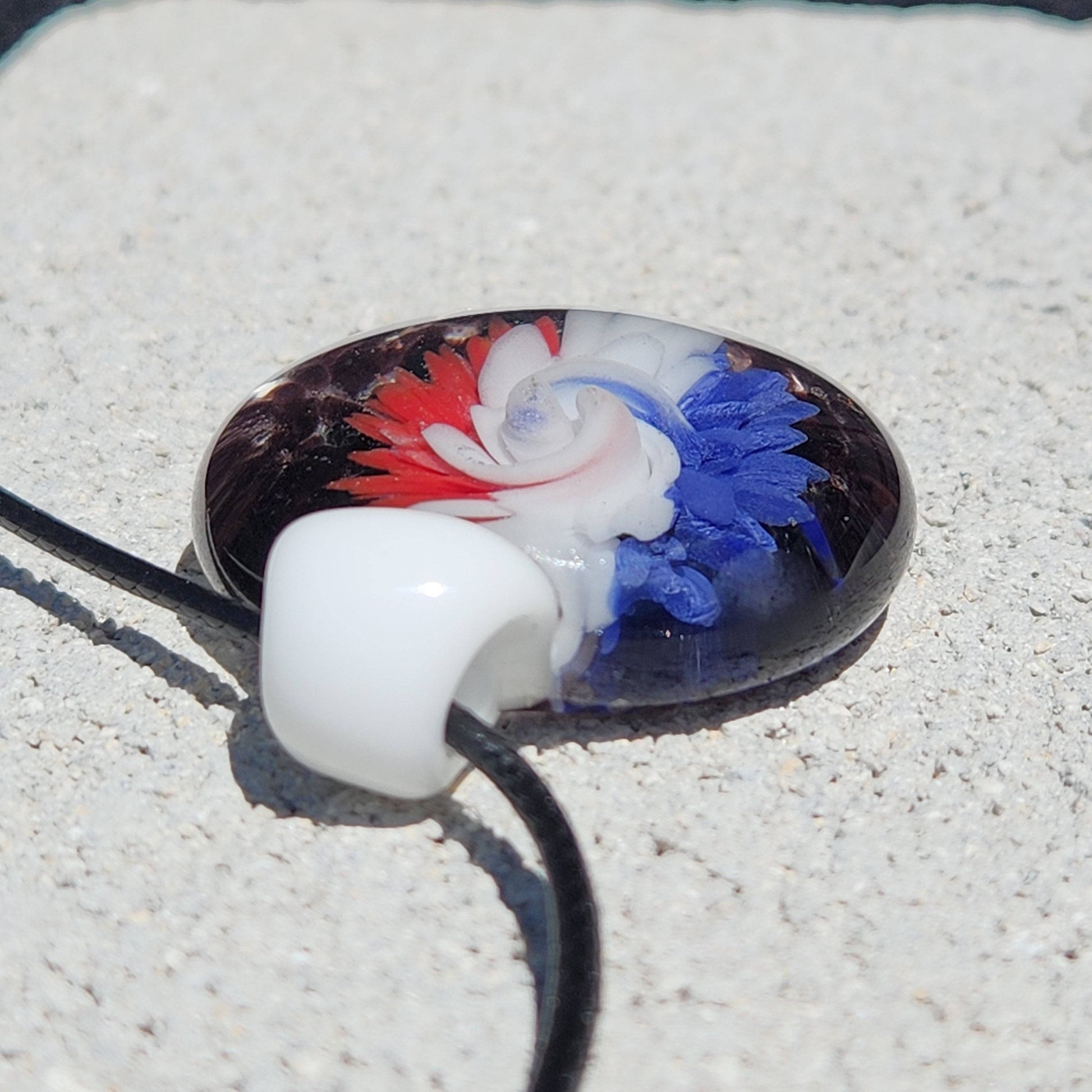 Artisan glass pendant with bold red, white, and blue flower swirl on black background. Handmade charm for stylish necklaces.