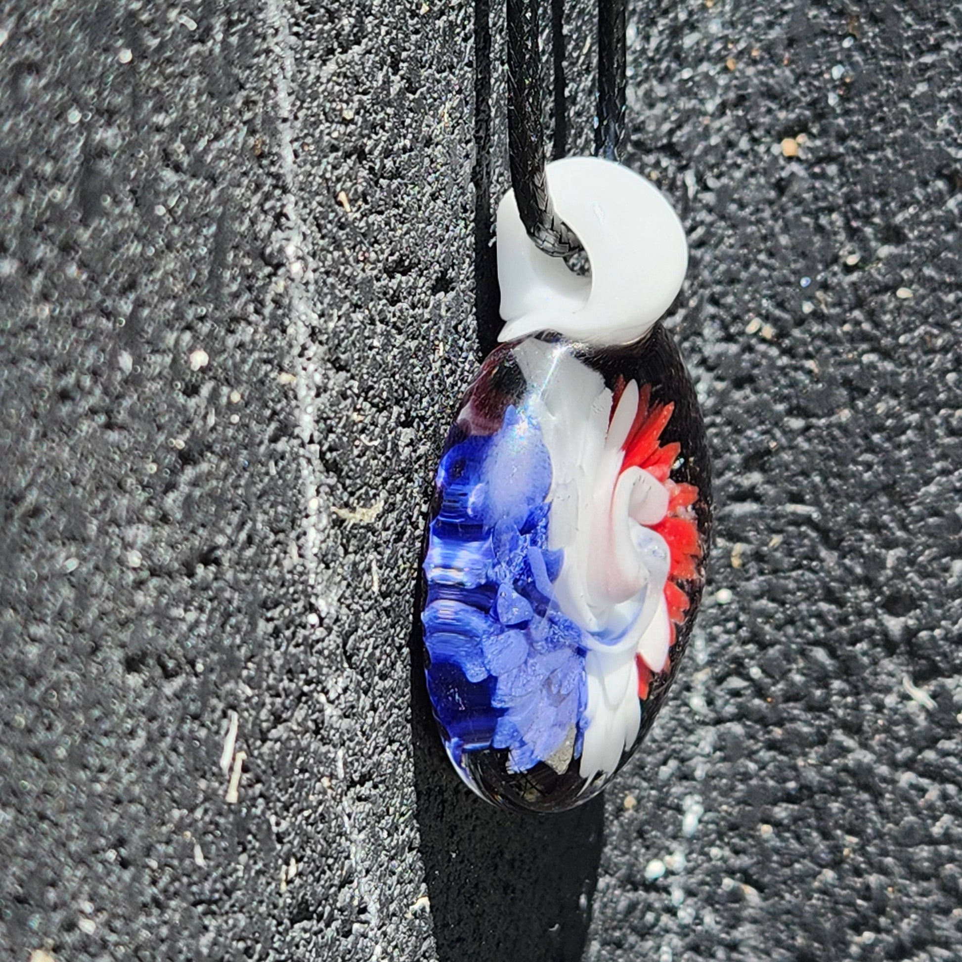 Vertical view of unique handmade glass pendant with red, white, and blue swirl. Trippy floral design on black borosilicate glass.