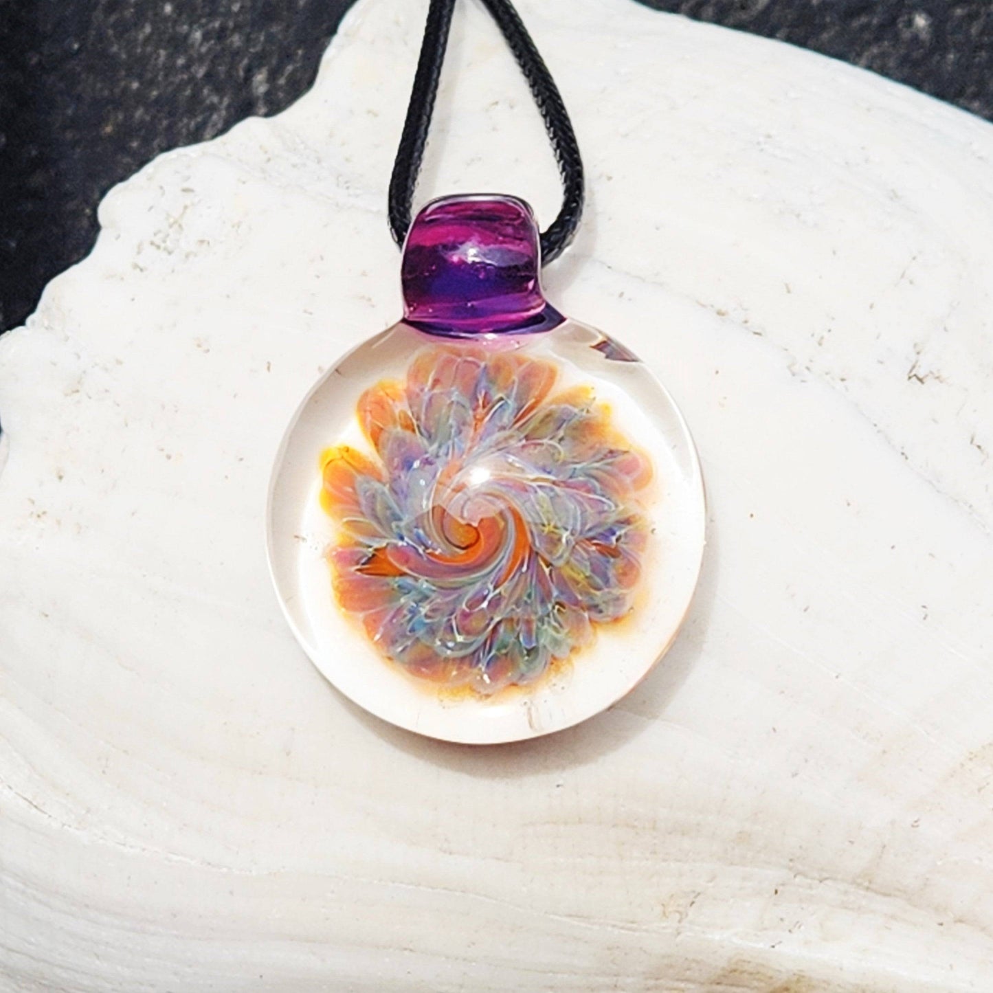 Small blown glass pendant statement necklace by DragonFire Glass featuring a swirling flower design in purples, amber, and blue on a black cord.