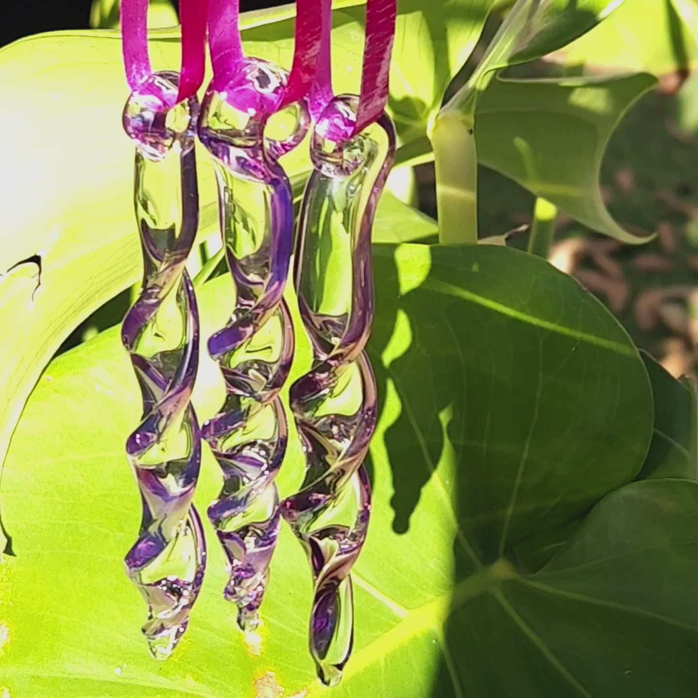 Three purple spiral glass suncatchers casting reflections on green foliage, enhancing garden ambiance with handmade charm.
