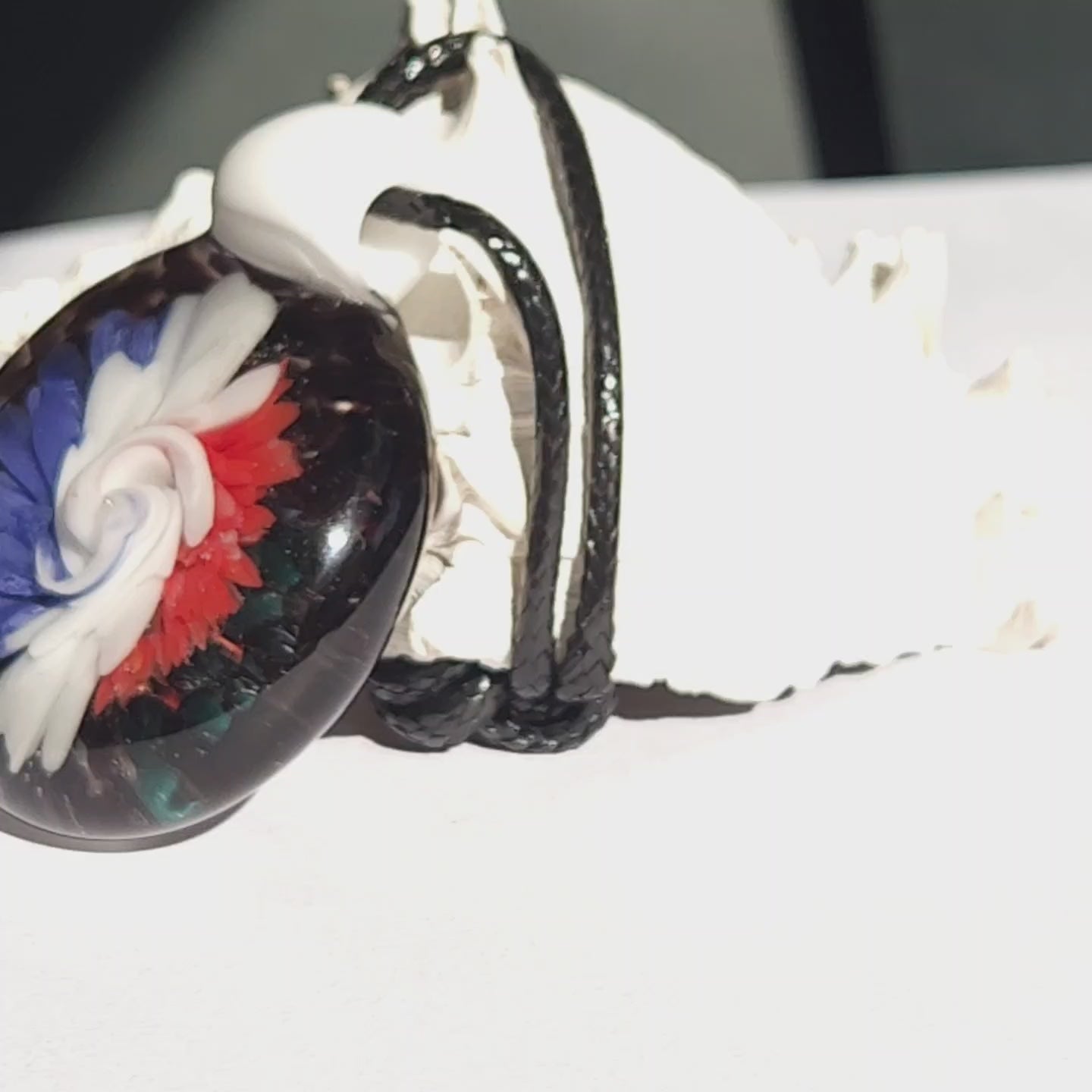 Close-up of trippy glass pendant necklace with vivid red, white, and blue swirls. Artistic handmade jewelry piece with black cord.
