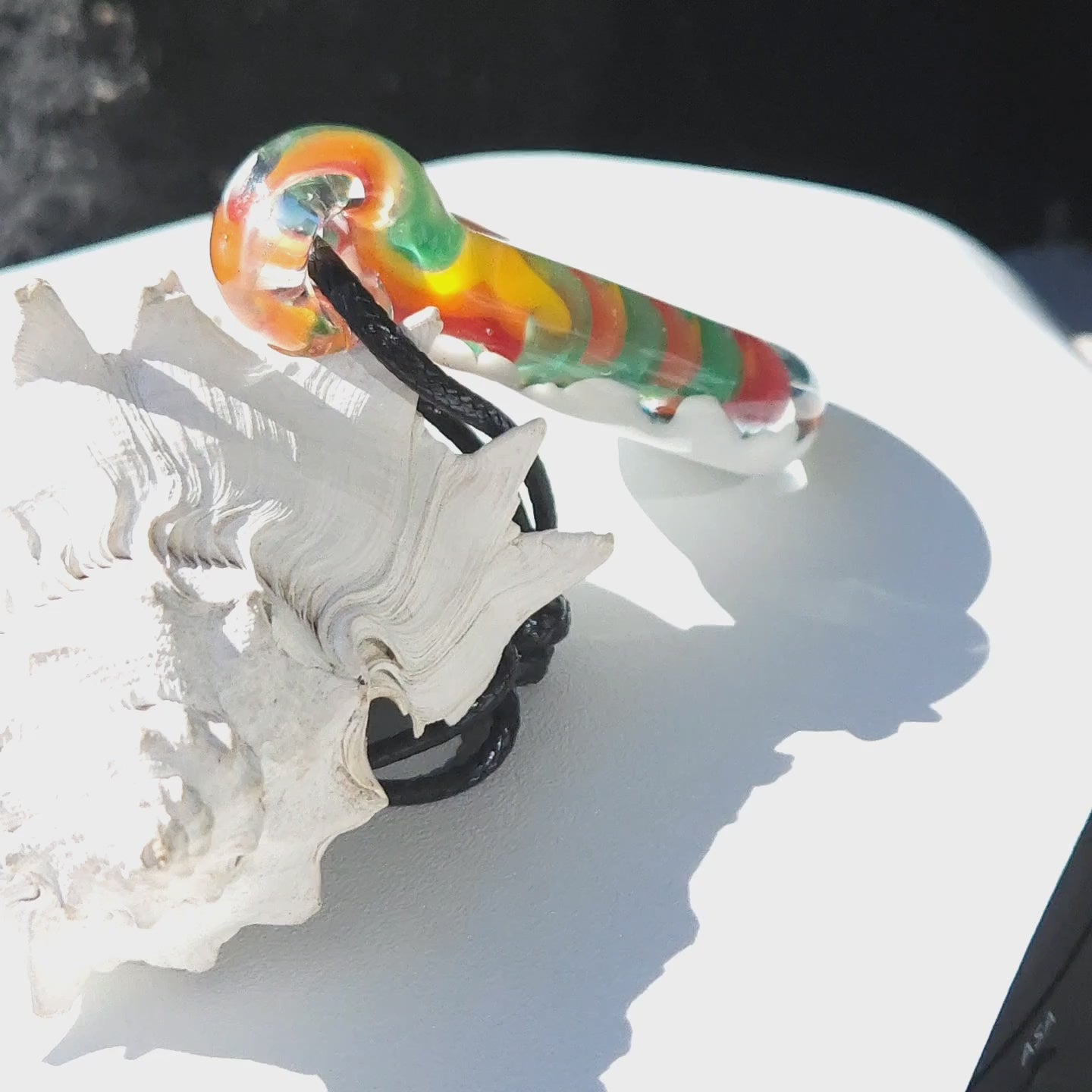 Side view of handmade glass pendant necklace showing rainbow swirl pattern and black cord, resting on a white seashell and surface.