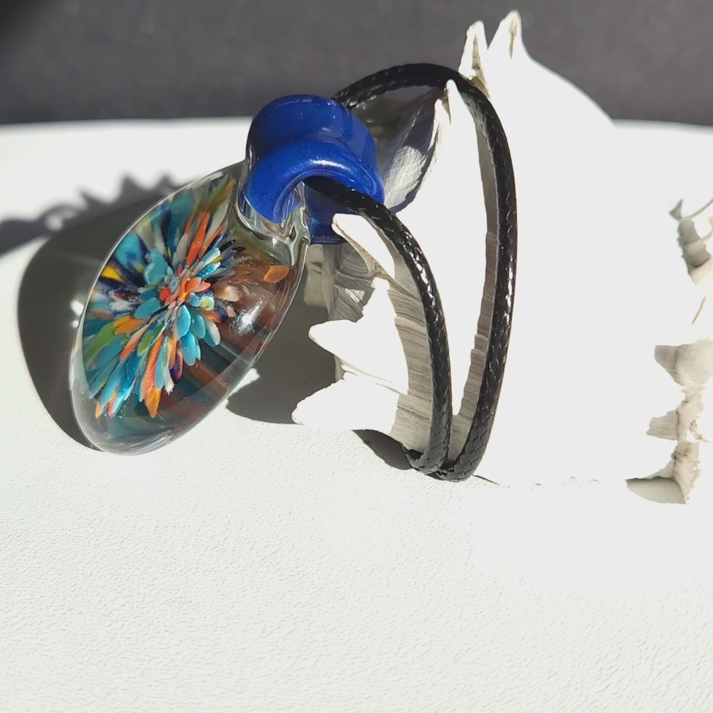 Trippy glass pendant necklace by DragonFire Glass featuring a multicolored floral burst inside clear glass with a bold blue hook on black cord.