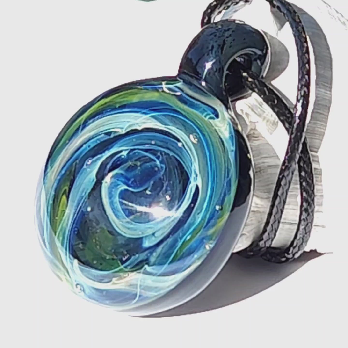 Detailed side angle of handmade borosilicate glass pendant with swirling blue and green galaxy pattern, suspended on a black cord.