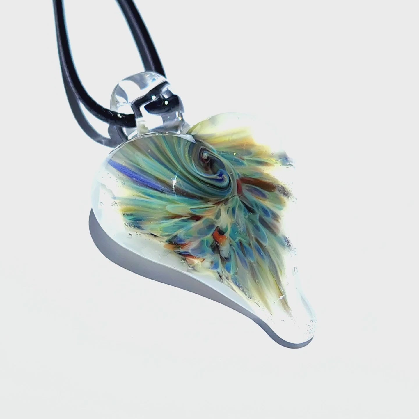 Heart-shaped handmade glass pendant with psychedelic swirl design in blue, green, and amber, suspended on a black cord against white background.