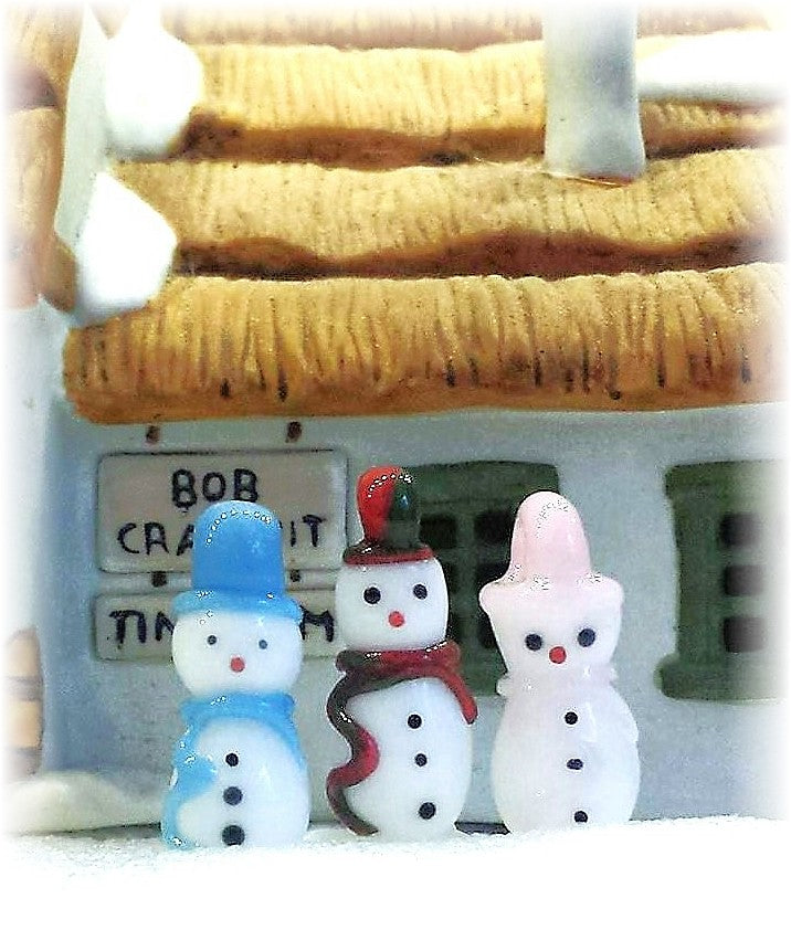 Colorful holiday glass snowman ornaments in front of a snowy cottage.