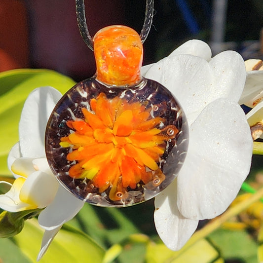 Handmade glass pendant necklace featuring fiery orange and yellow floral swirl, hanging in front of white tropical flowers.