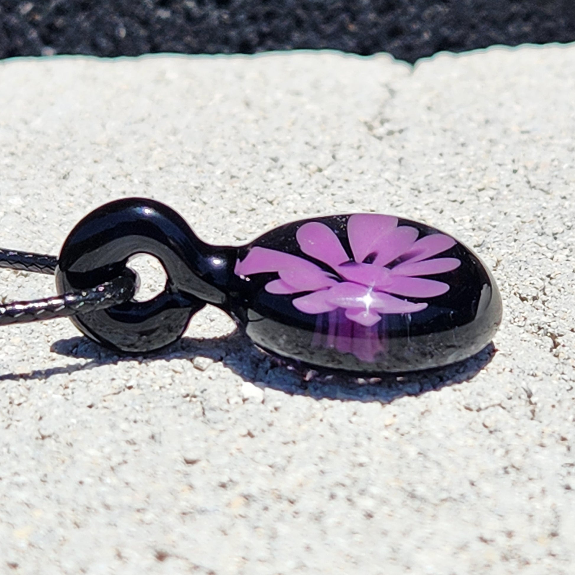 Close-up of small handmade purple flower glass pendant on black background, ideal for unique gift jewelry, lying flat on stone surface.