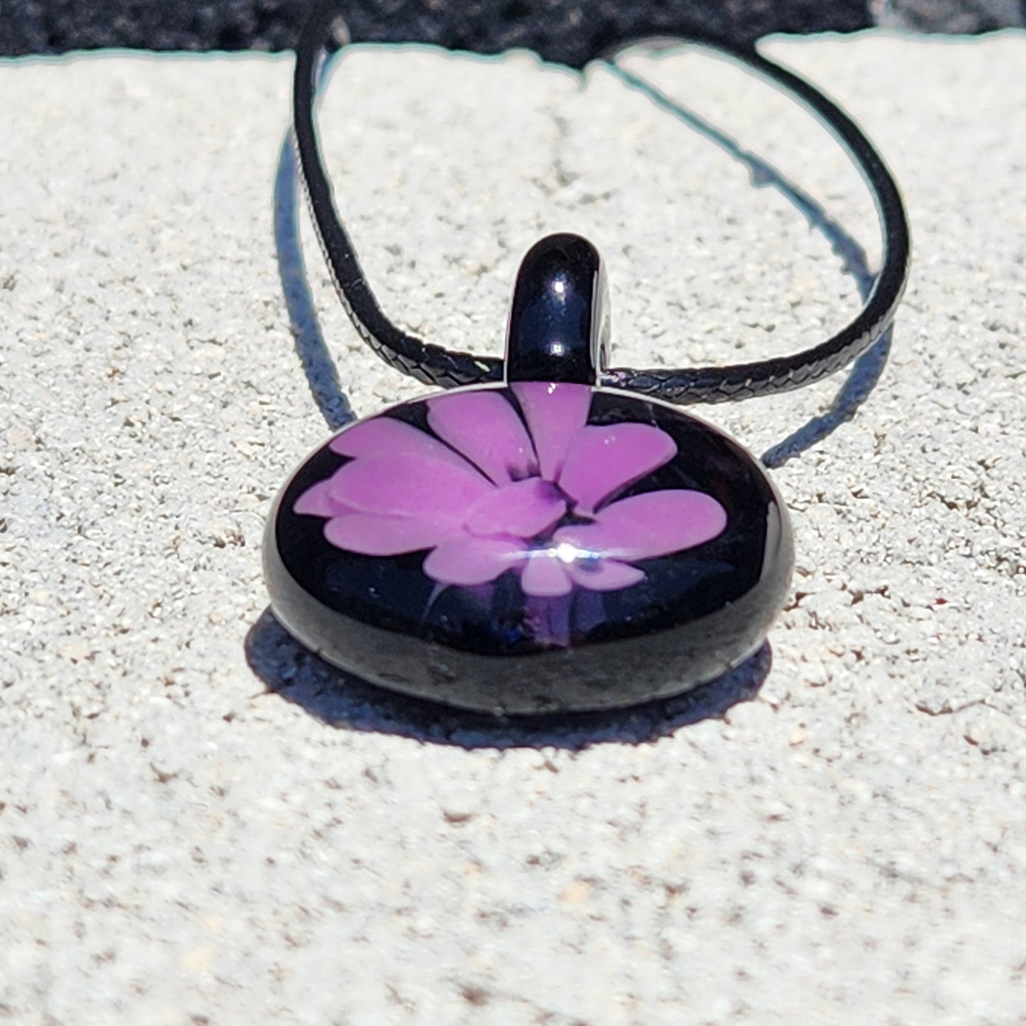 Front view of small handmade purple flower glass pendant on black cord, minimalist jewelry piece photographed on concrete surface.