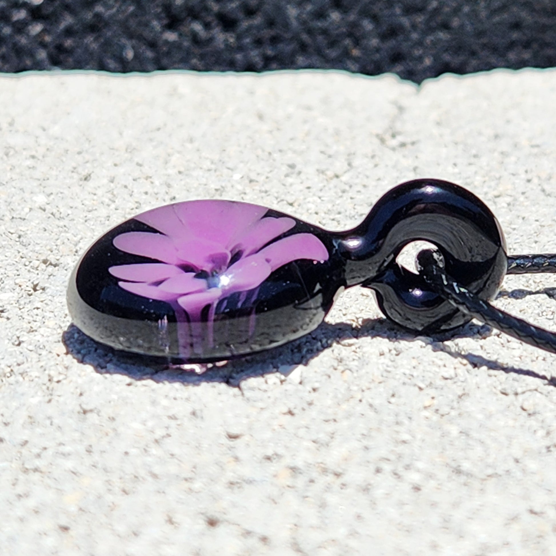 Side angle of minimalist purple and black glass pendant necklace, handmade in the USA, resting on stone surface for product detail view.
