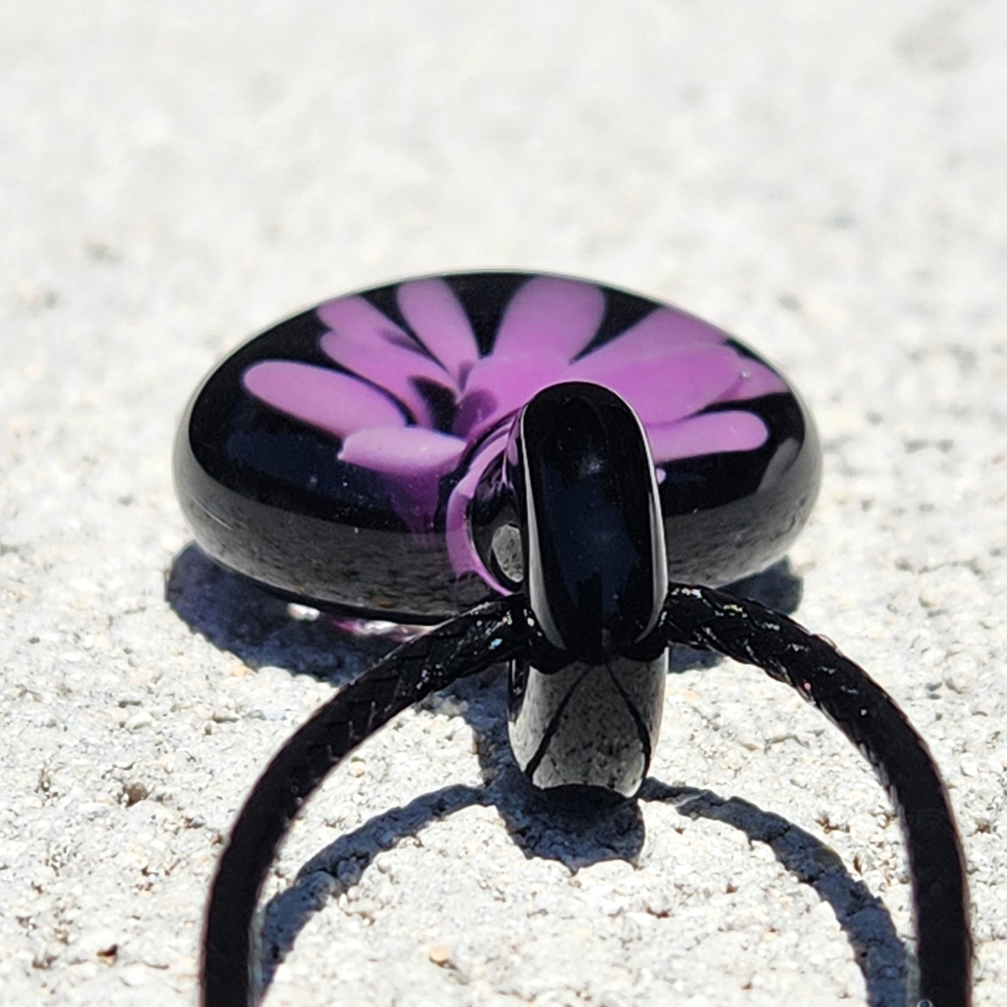 Bottom view of minimalist purple flower glass pendant with black loop, handmade in the USA, displayed on light stone surface.