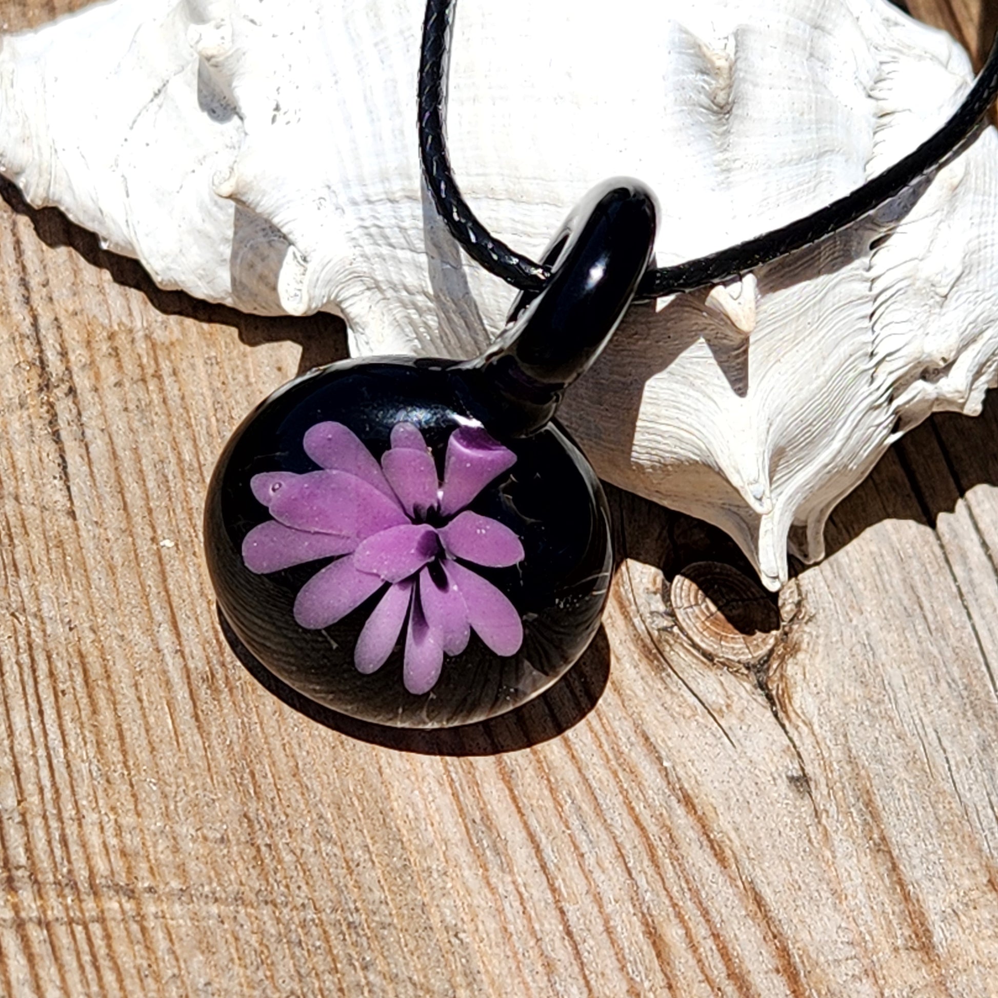 Small handmade purple flower pendant on black cord, displayed on wood with shell, ideal for minimalist jewelry and gift ideas.