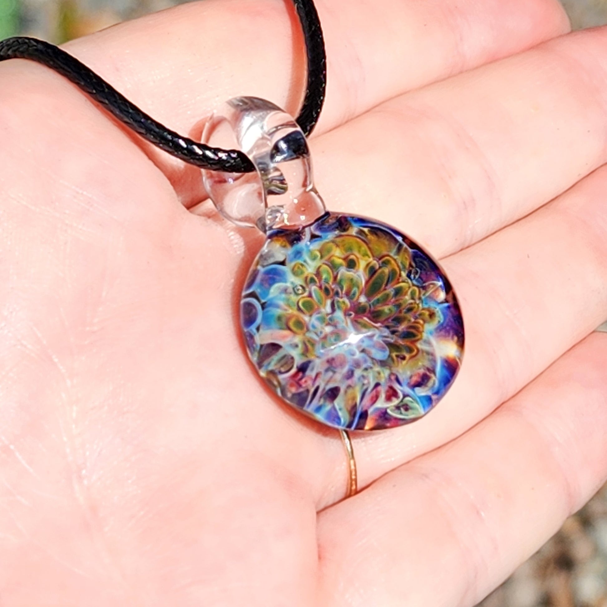 Hand holding Ocean Reef Glass Pendant Necklace with vivid reef-inspired glasswork in blue, green, and red tones under sunlight.