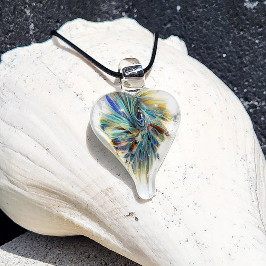 Handmade blown glass pendant in heart shape with trippy blue, green, and white swirl design, displayed on a white seashell background.