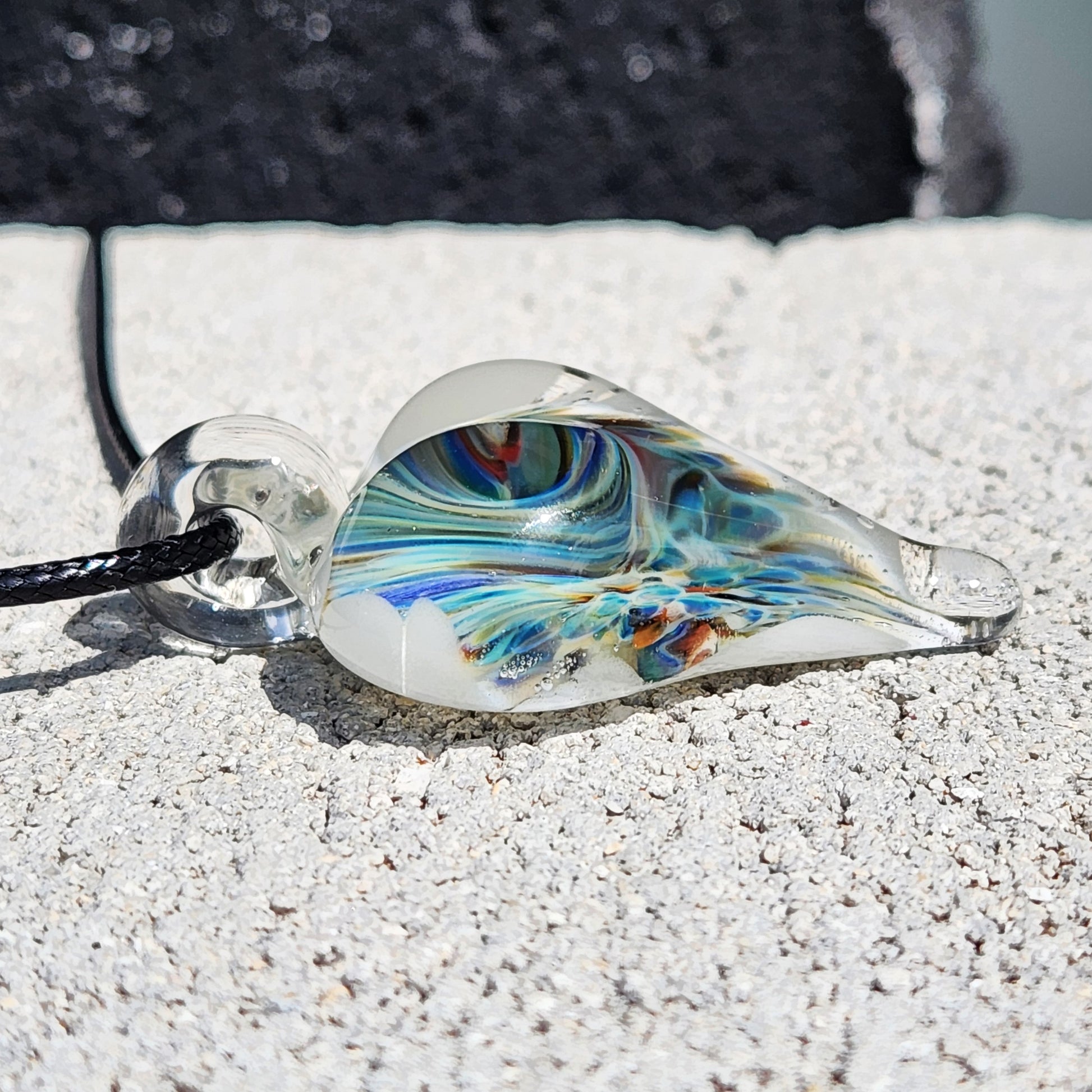 Side profile of handmade blown glass heart pendant with swirling blue and green design, lying flat on a textured stone surface.