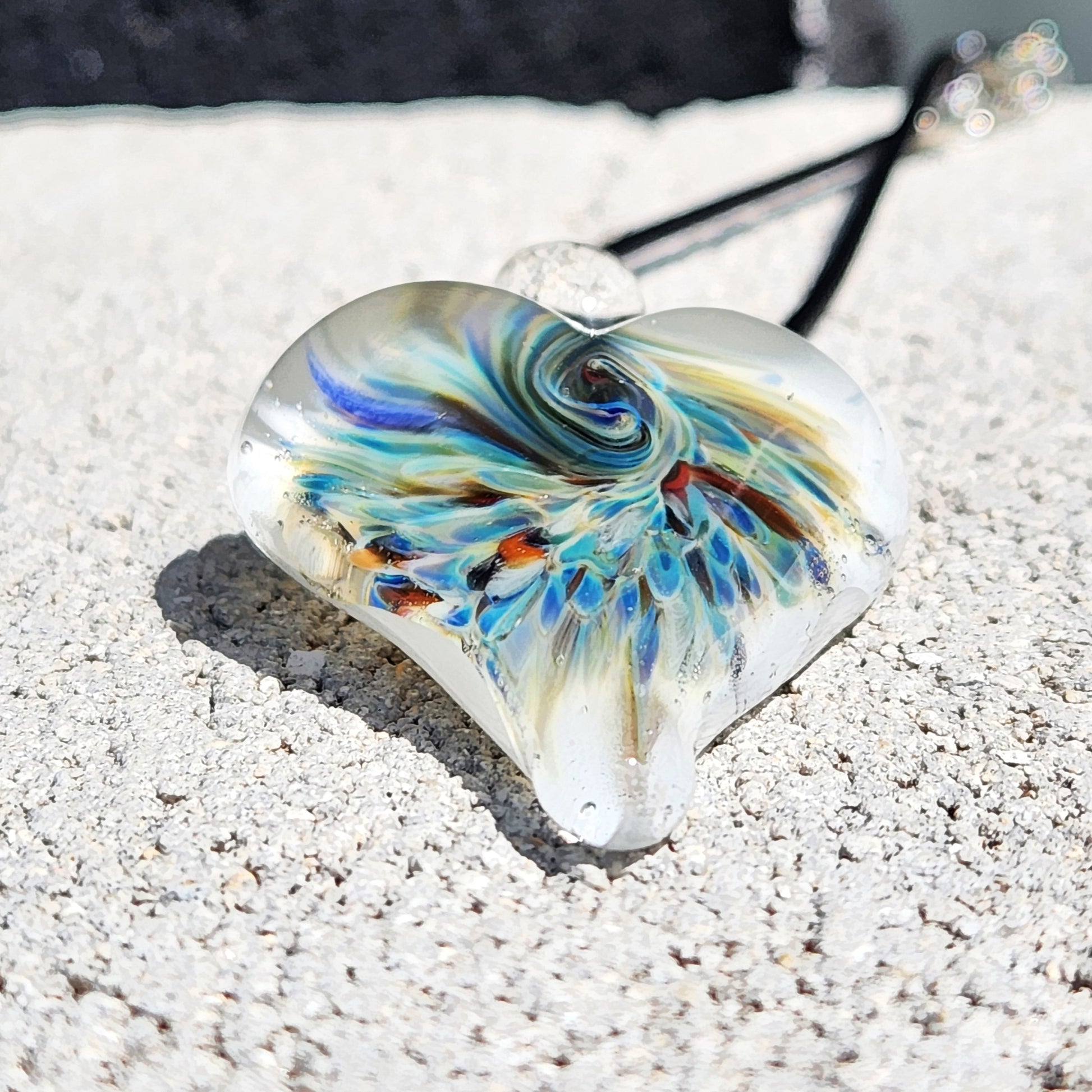 Heart-shaped handmade glass pendant featuring a vibrant implosion design in blue and green tones, resting on a textured stone surface.