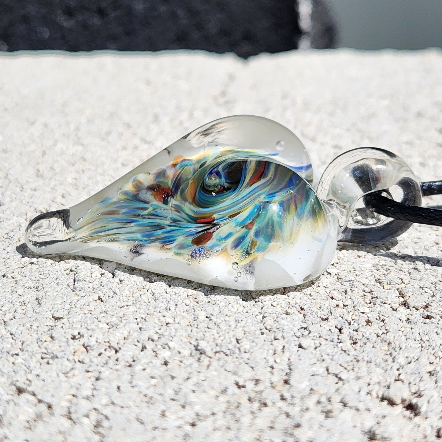 Detailed view of heart-shaped blown glass pendant with unique trippy implosion design in blue, green, and amber hues.