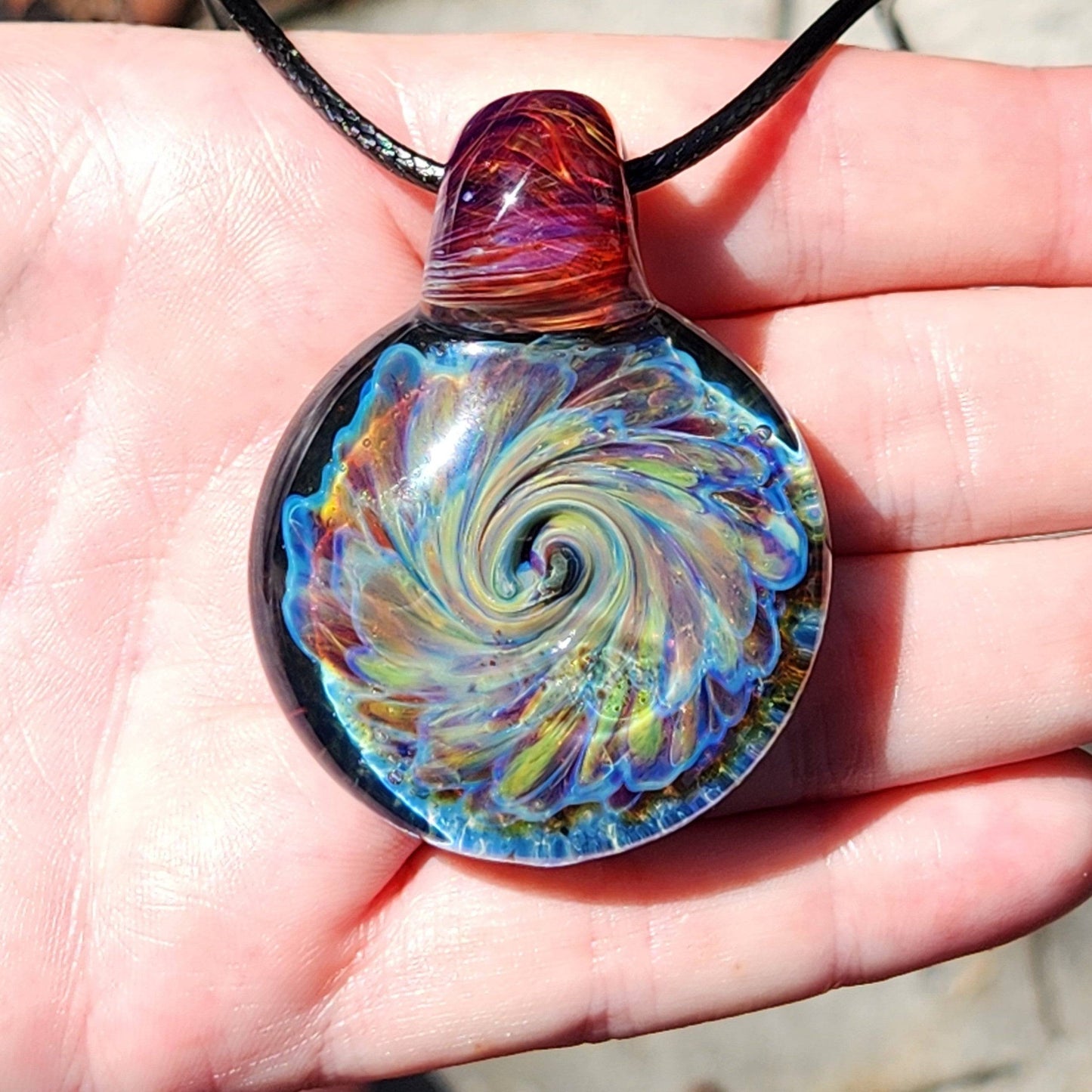 Multicolor glass pendant necklace with swirling design and red-amber loop, shown in hand for size and detail view.