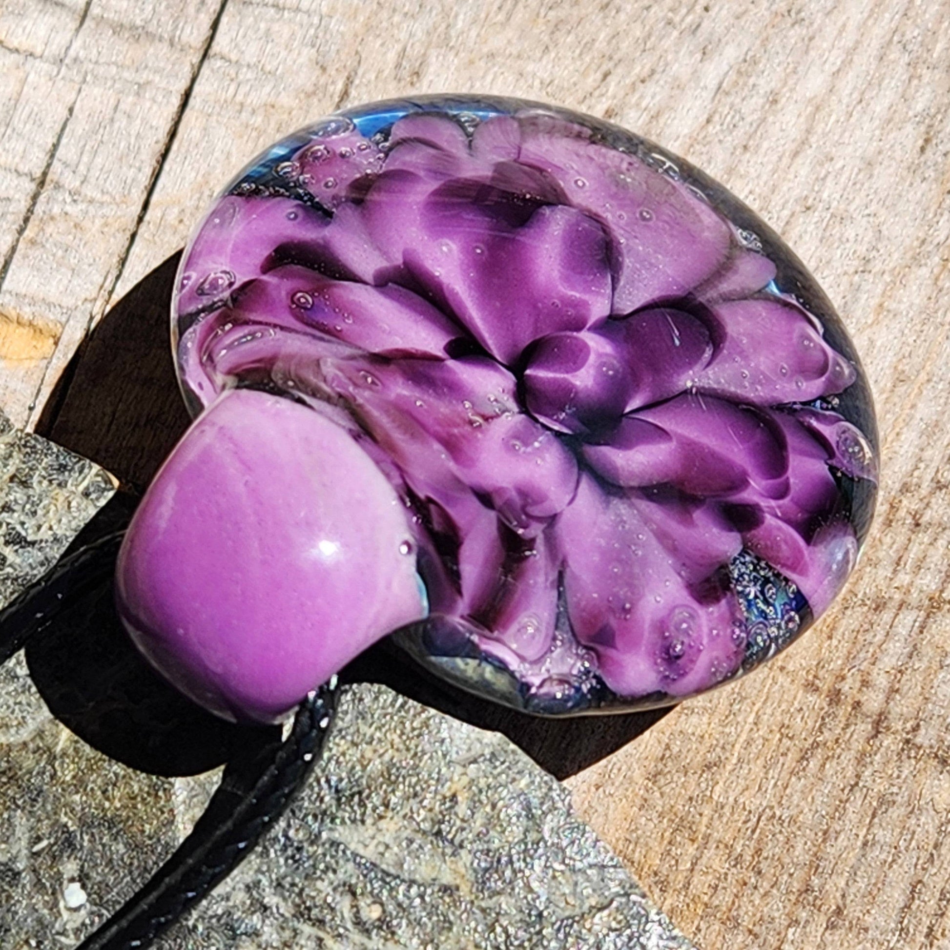 Artistic glass pendant with blooming purple flower design, resting on wood. Handcrafted borosilicate jewelry for unique style statements.