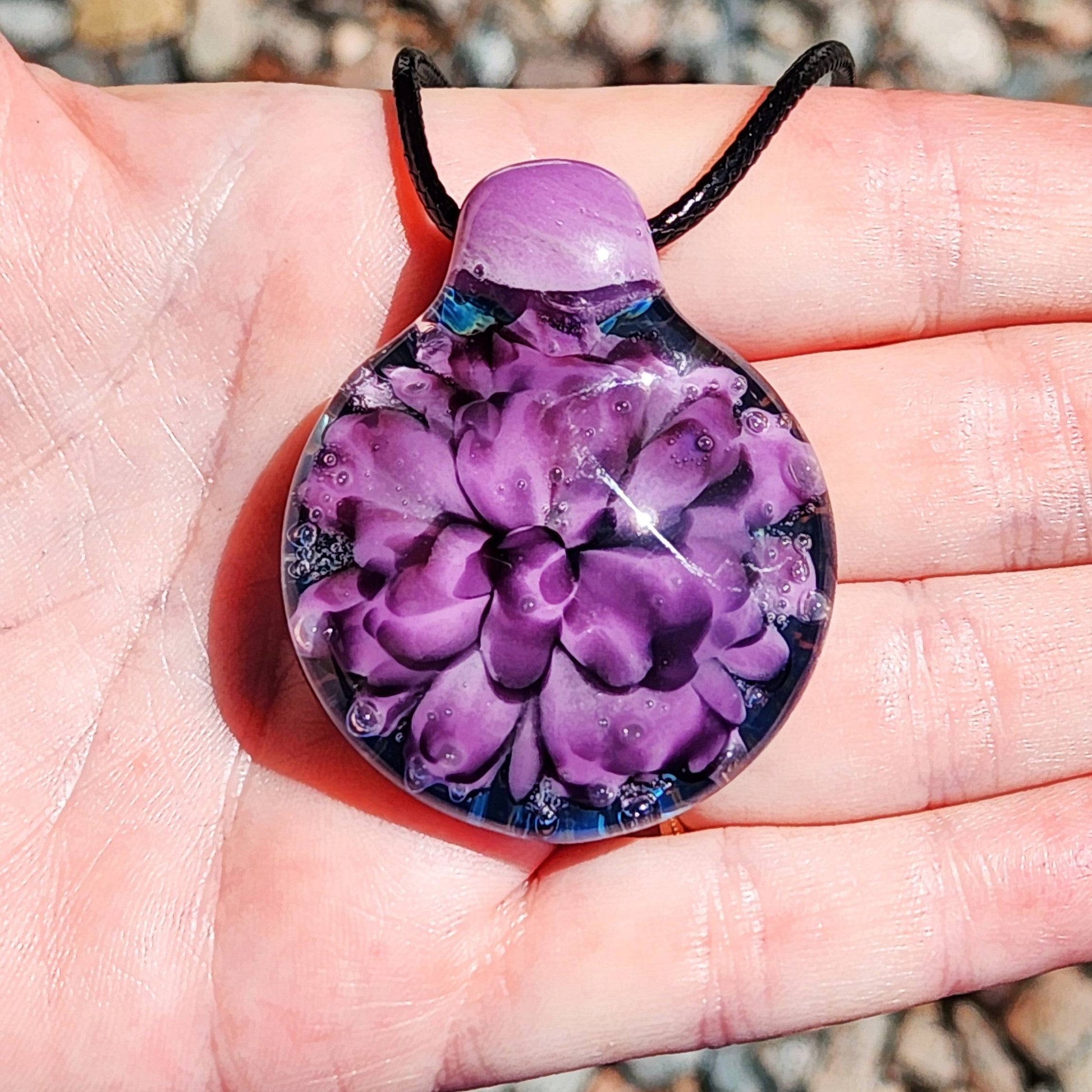 Hand holding a purple and black floral glass pendant necklace, showing size and detail. Unique borosilicate jewelry for everyday wear.