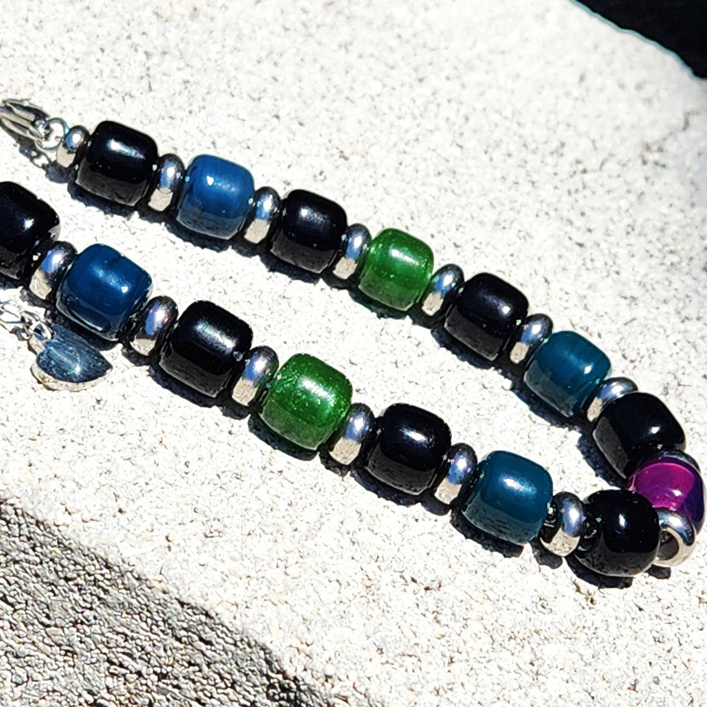 Pair of Boho Glass Beaded Bracelets with black, green, blue, and purple beads laid on a stone surface under sunlight.
