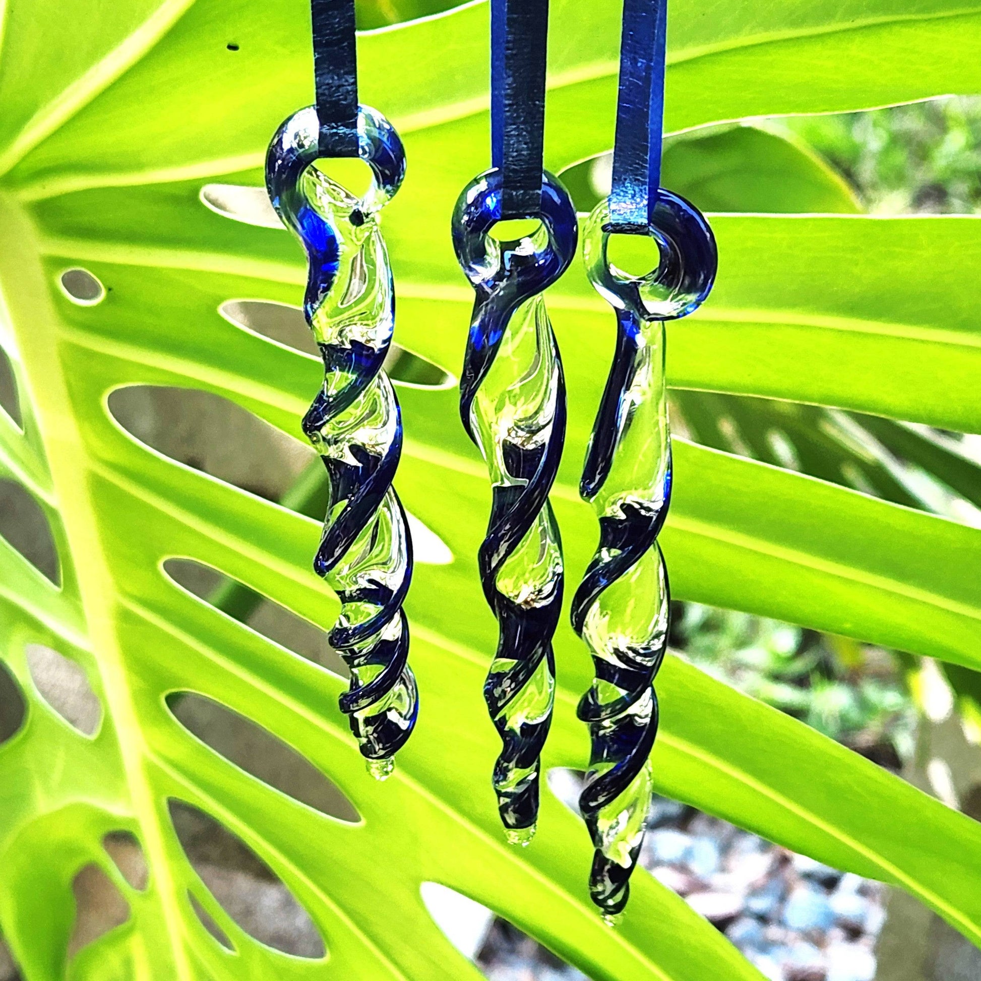 Three handmade blue glass icicle ornaments hanging on a tropical leaf. Unique Christmas suncatchers for indoor or outdoor holiday décor.
