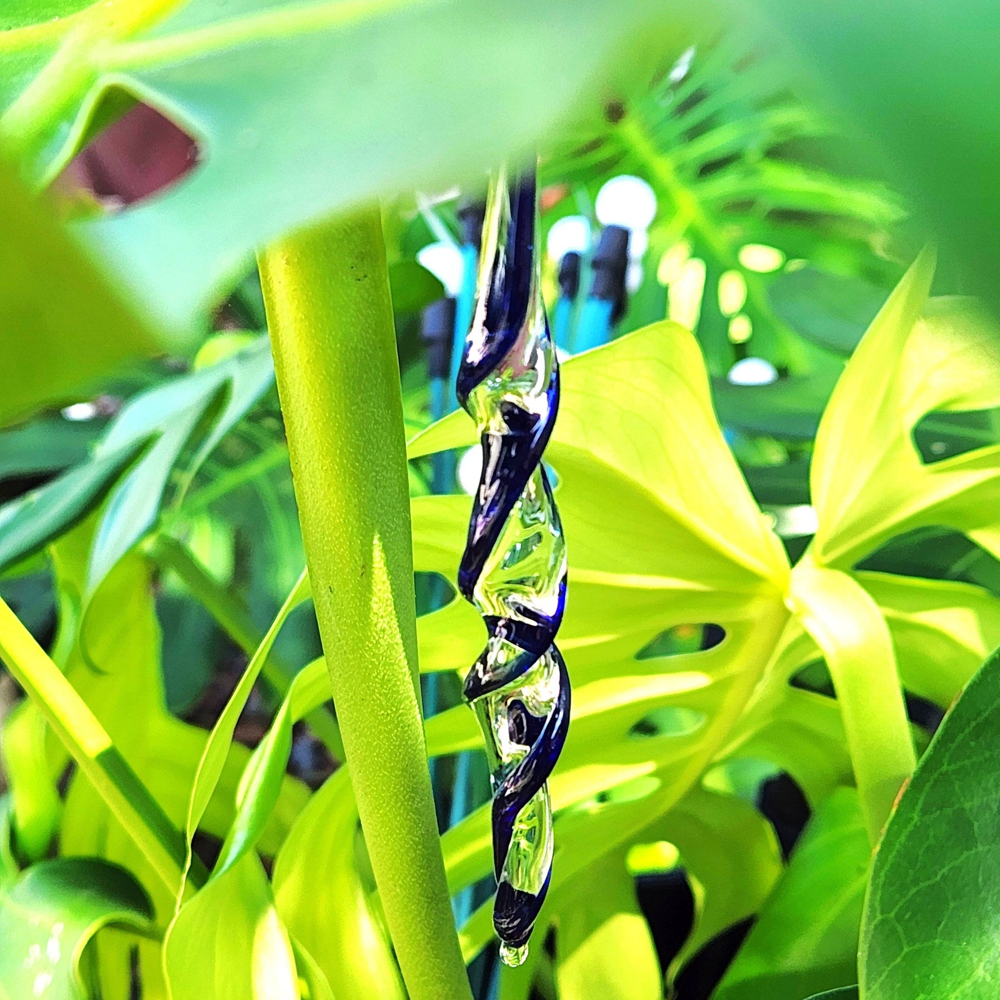 Single handmade blue glass icicle ornament hanging among green leaves. Artistic holiday suncatcher for garden or tree décor.