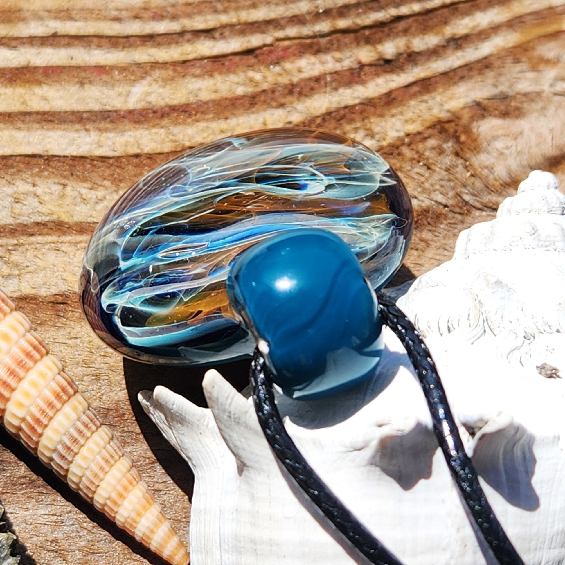 Close-up of handmade borosilicate glass pendant with gold, silver, and blue swirls. Unique jewelry gift resting on seashells and wood surface.