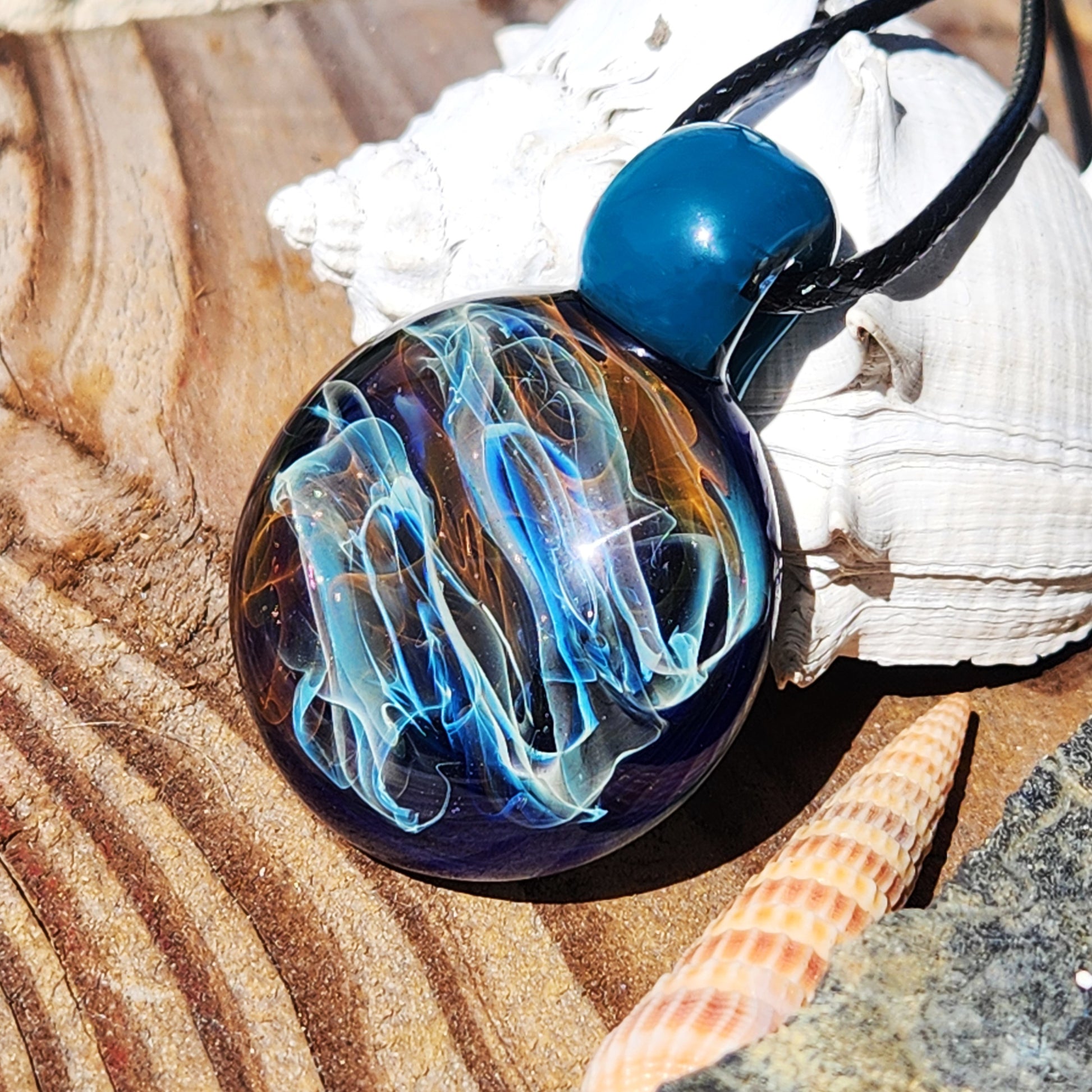 Handmade blown glass pendant necklace with swirling gold, silver, and blue hues on a black cord. Unique borosilicate jewelry gift on wood and shell.