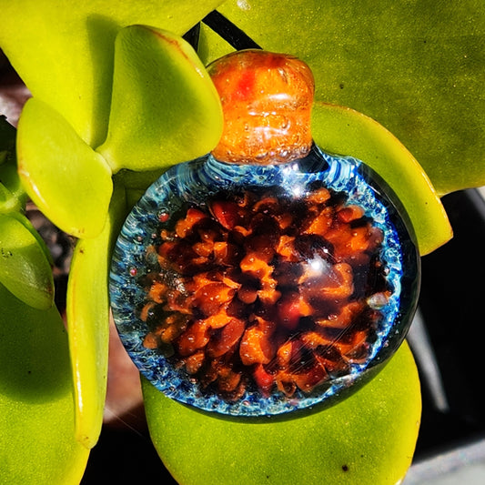 Handmade glass pendant with crushed borosilicate in fiery orange and blue hues, displayed on green succulent leaves. Unique gift-ready jewelry.