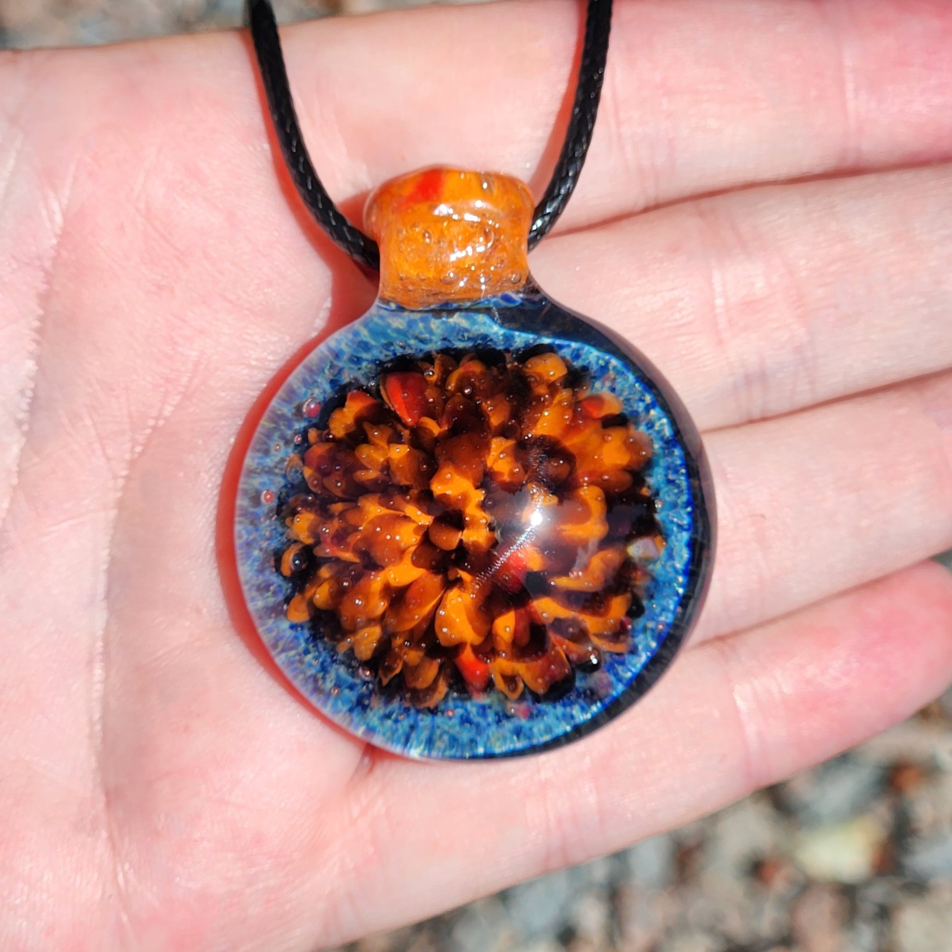 Handheld view of handmade glass pendant with fiery orange and blue crushed borosilicate, highlighting its vibrant colors and compact size.