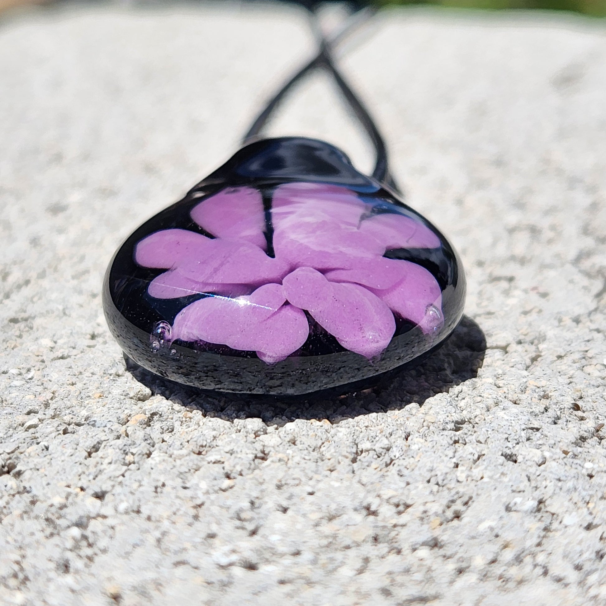 Close-up of small handmade purple flower glass pendant on black cord, showcasing glossy finish and intricate petal details.