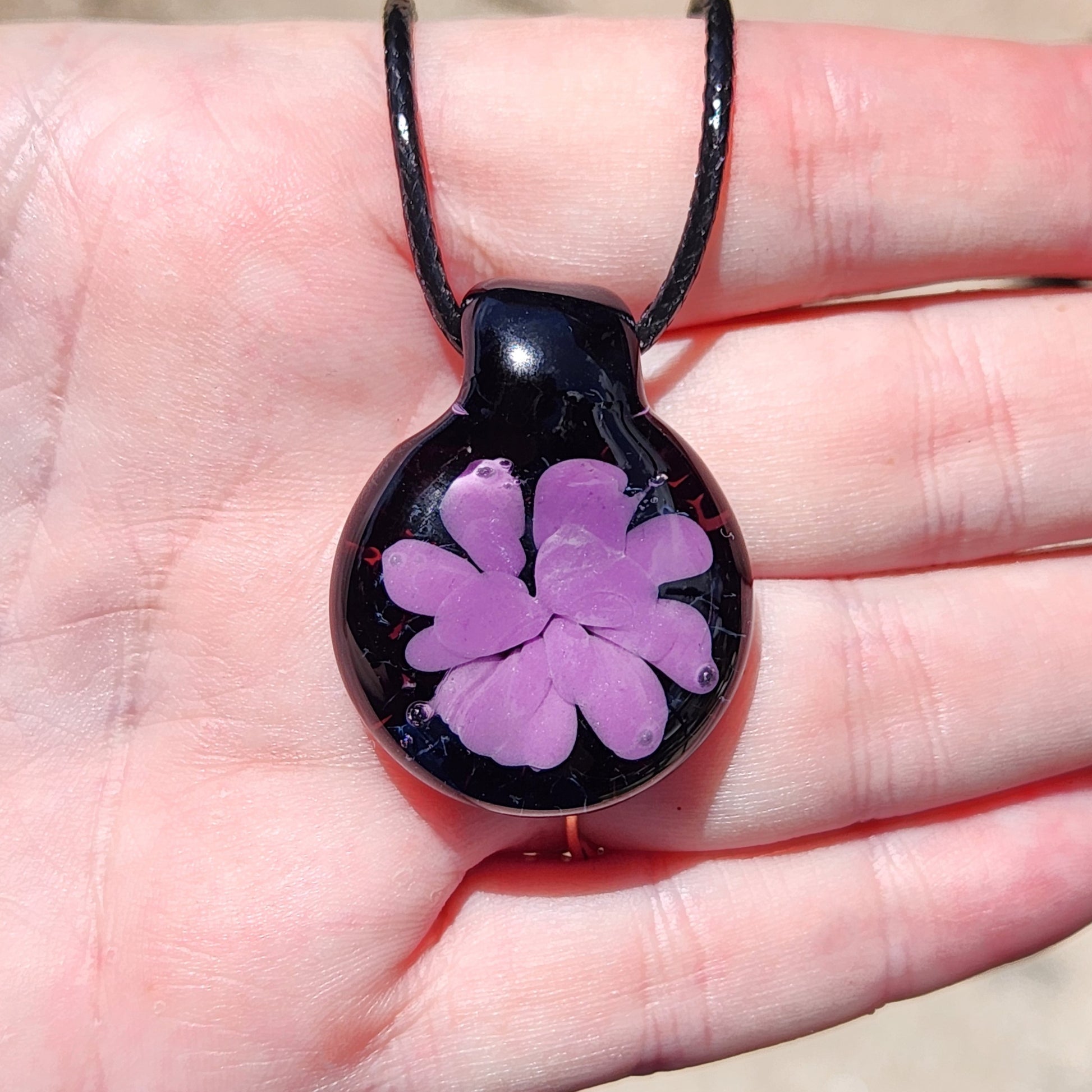 Handheld view of minimalist purple flower glass pendant on black cord, showcasing its size and elegant handmade detail.