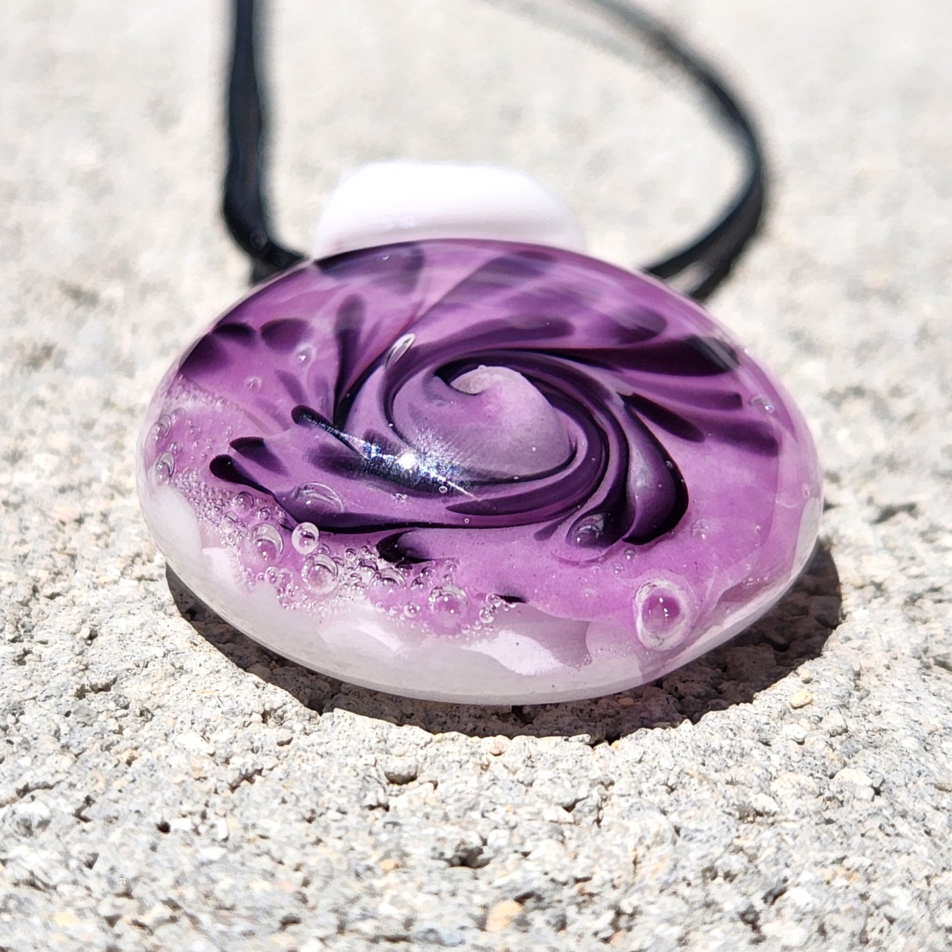 Close-up of handmade purple spiral glass pendant necklace with black accents and white base, shown on a concrete background.