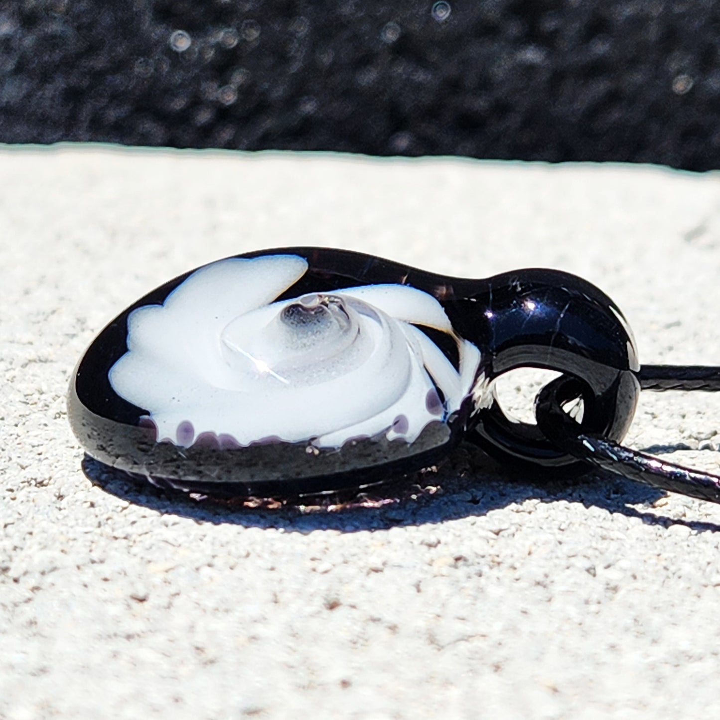 Side angle of handmade black and white swirl glass pendant necklace showing its curved shape and layered swirl design on textured surface.