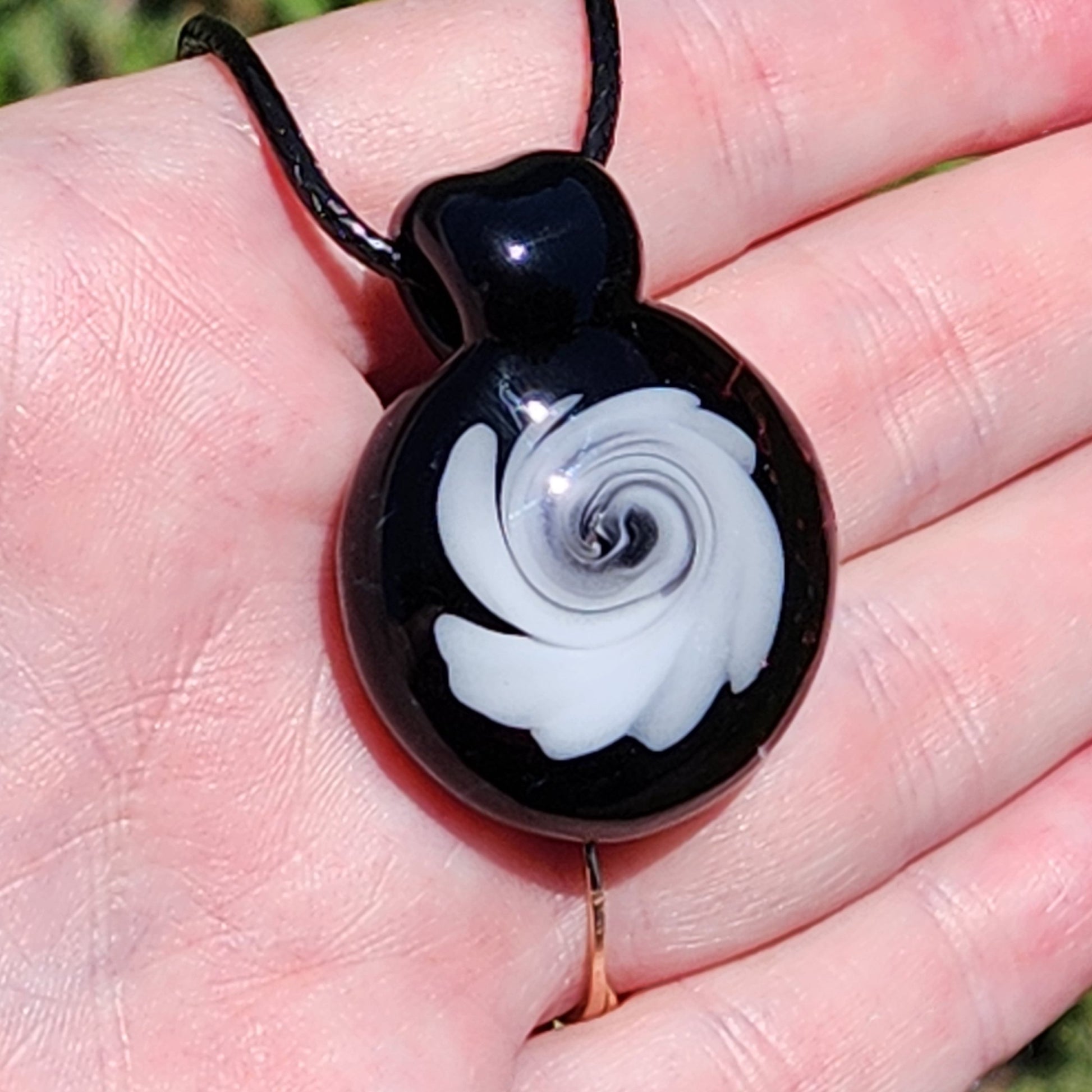 Hand holding handmade black and white swirl glass pendant necklace outdoors, displaying its compact size and elegant swirl pattern in sunlight.
