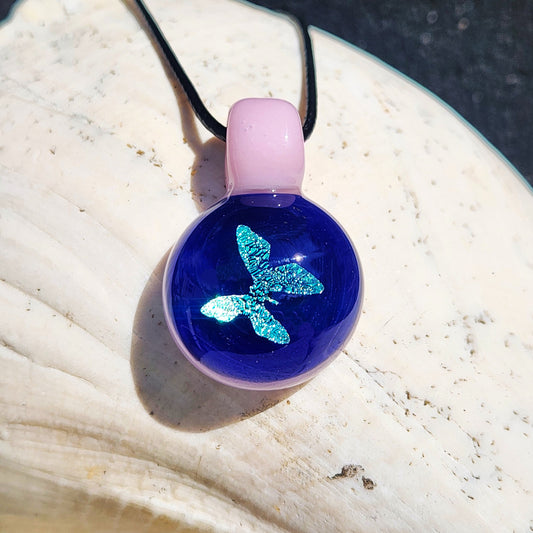 Small glass butterfly pendant with shimmering blue dichroic design and pink loop, displayed on a seashell. Handmade jewelry gift.