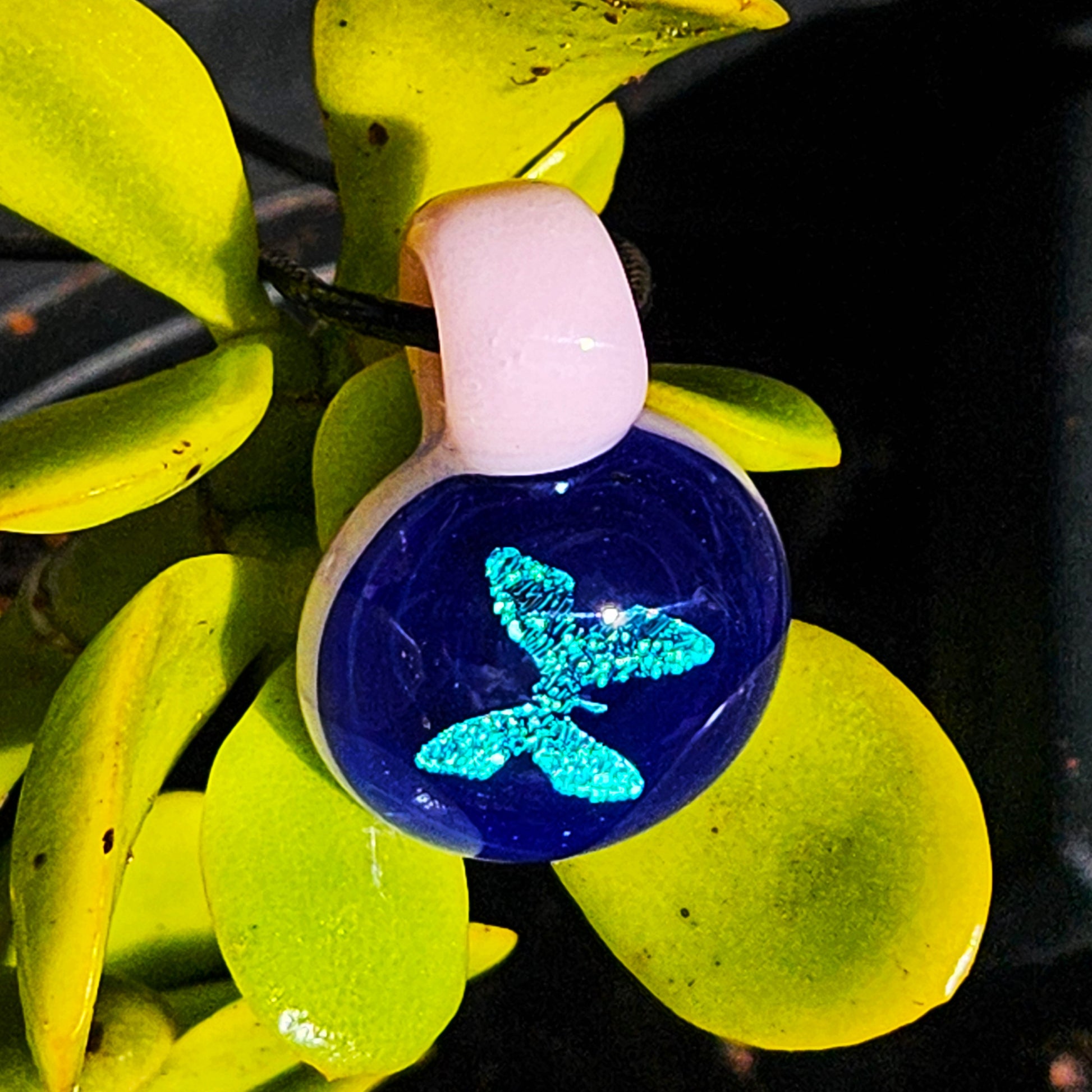 Blue dichroic butterfly pendant with pink loop hanging on a green plant, highlighting its vibrant handmade glass design.