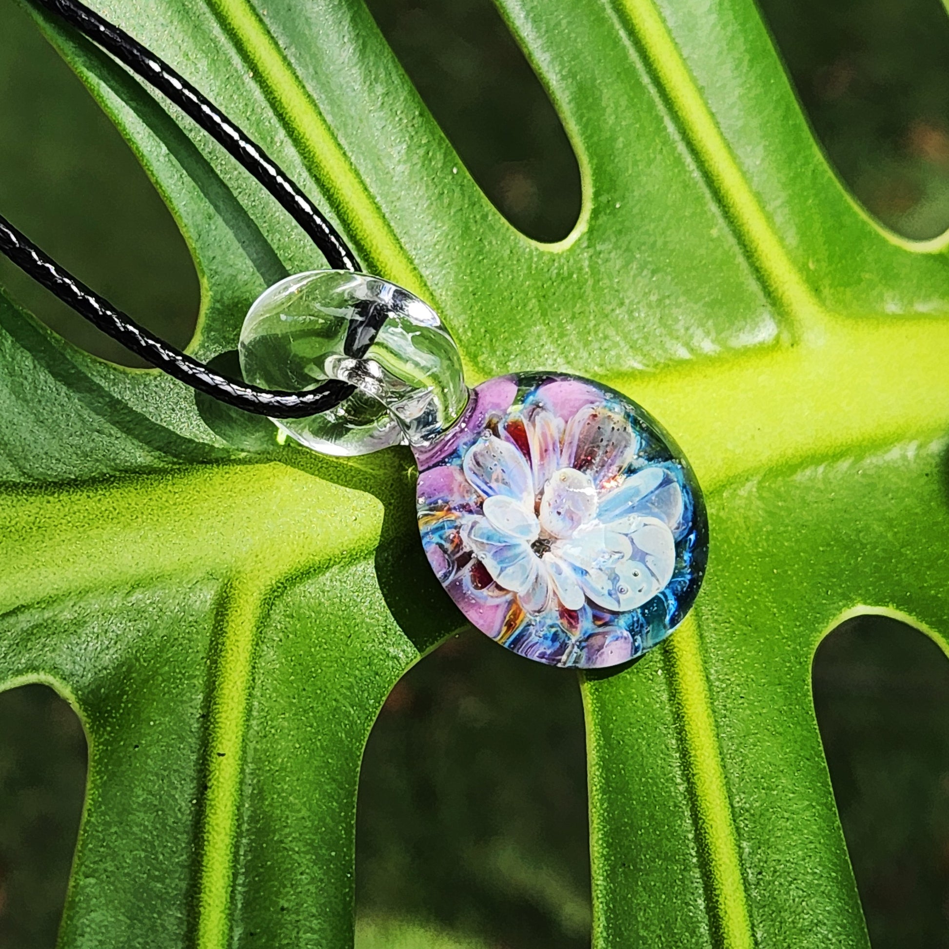 Handmade Borosilicate Glass Pendant on green leaf, featuring vibrant blue and purple bloom with UV-reactive design.