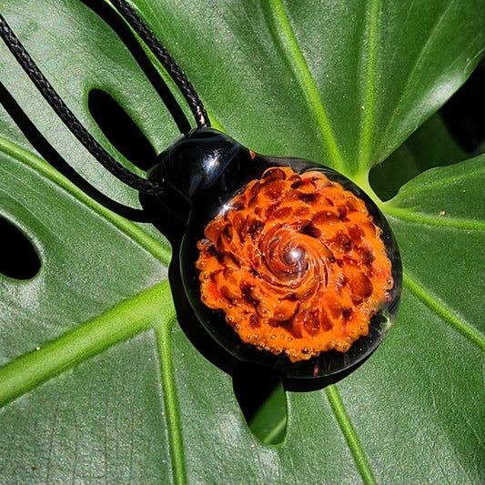 Handmade glass pendant necklace with bold orange and black spiral design displayed on a green monstera leaf. Unique boho jewelry gift.