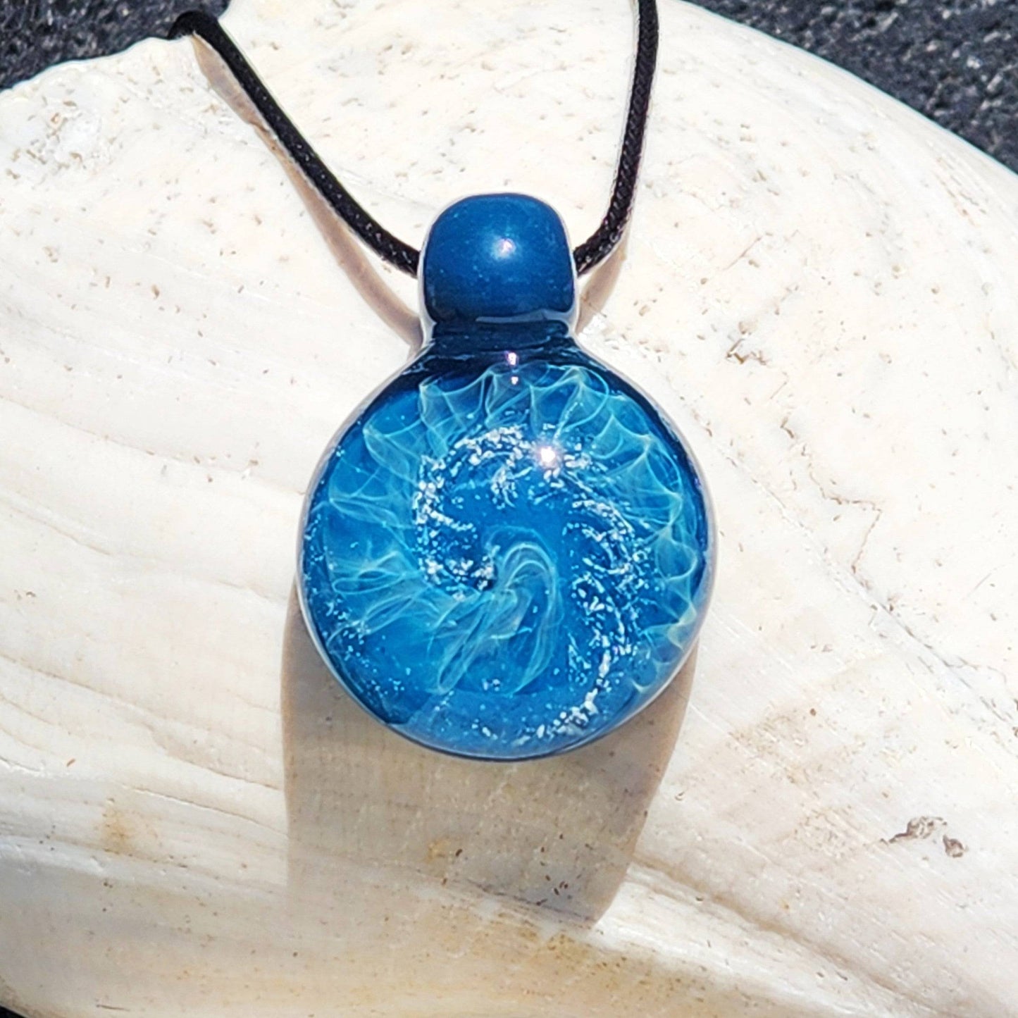 Handmade blown glass pendant necklace with swirling ocean-inspired design and silver highlights, displayed on a white seashell background.