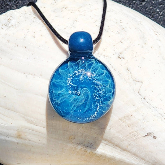 Handmade blown glass pendant necklace with swirling ocean-inspired design and silver highlights, displayed on a white seashell background.