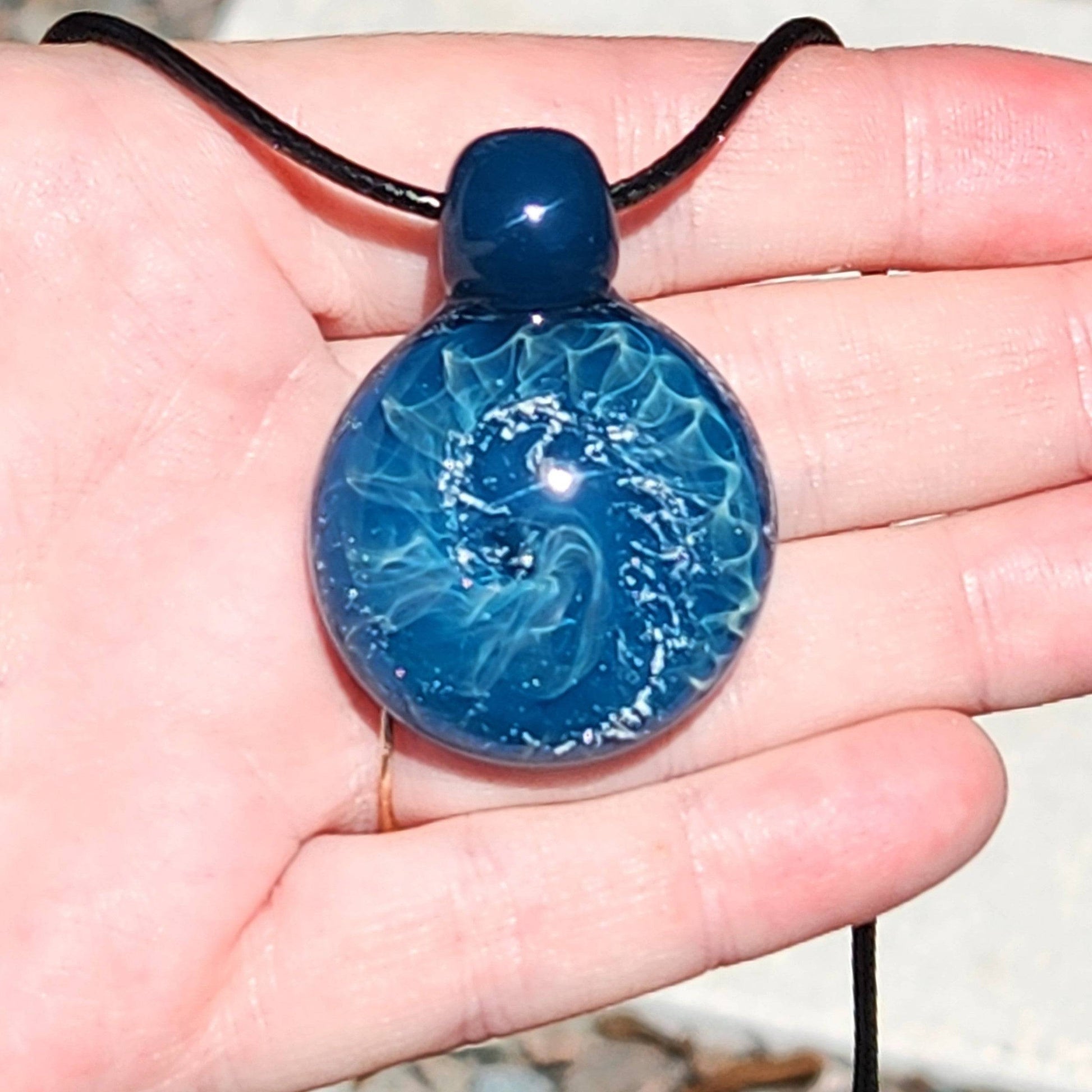 Ocean-inspired borosilicate glass pendant necklace with swirling wave design, displayed in hand for scale and detail.