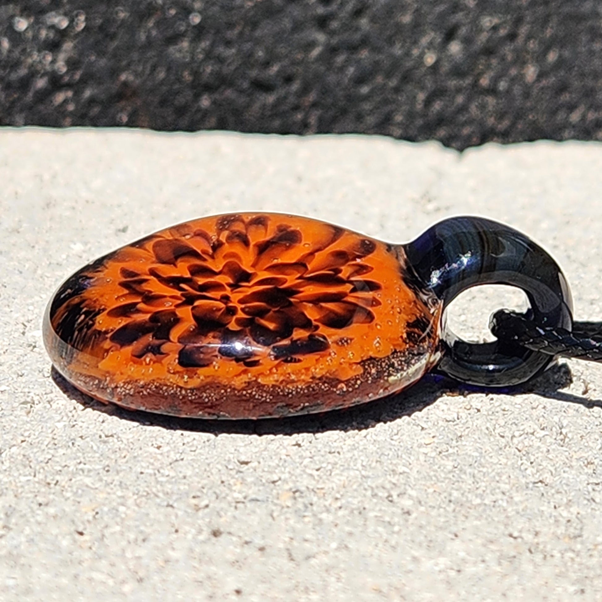 Fiery Flame Glass Pendant lying flat, showing intricate orange bloom and black glass loop. Handmade fall accessory for Halloween style.